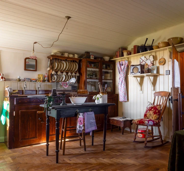 Cozy vintage kitchen at Boloquoy Farm & Watermill with dark wood cabinets, open shelving, a small wooden table, and a floral cushioned rocking chair.