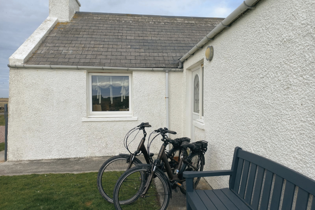 Two bicycles are parked outside near a white textured house with a small window and white door, with a dark blue bench on the right side. The house has a sloped roof with asphalt shingles.