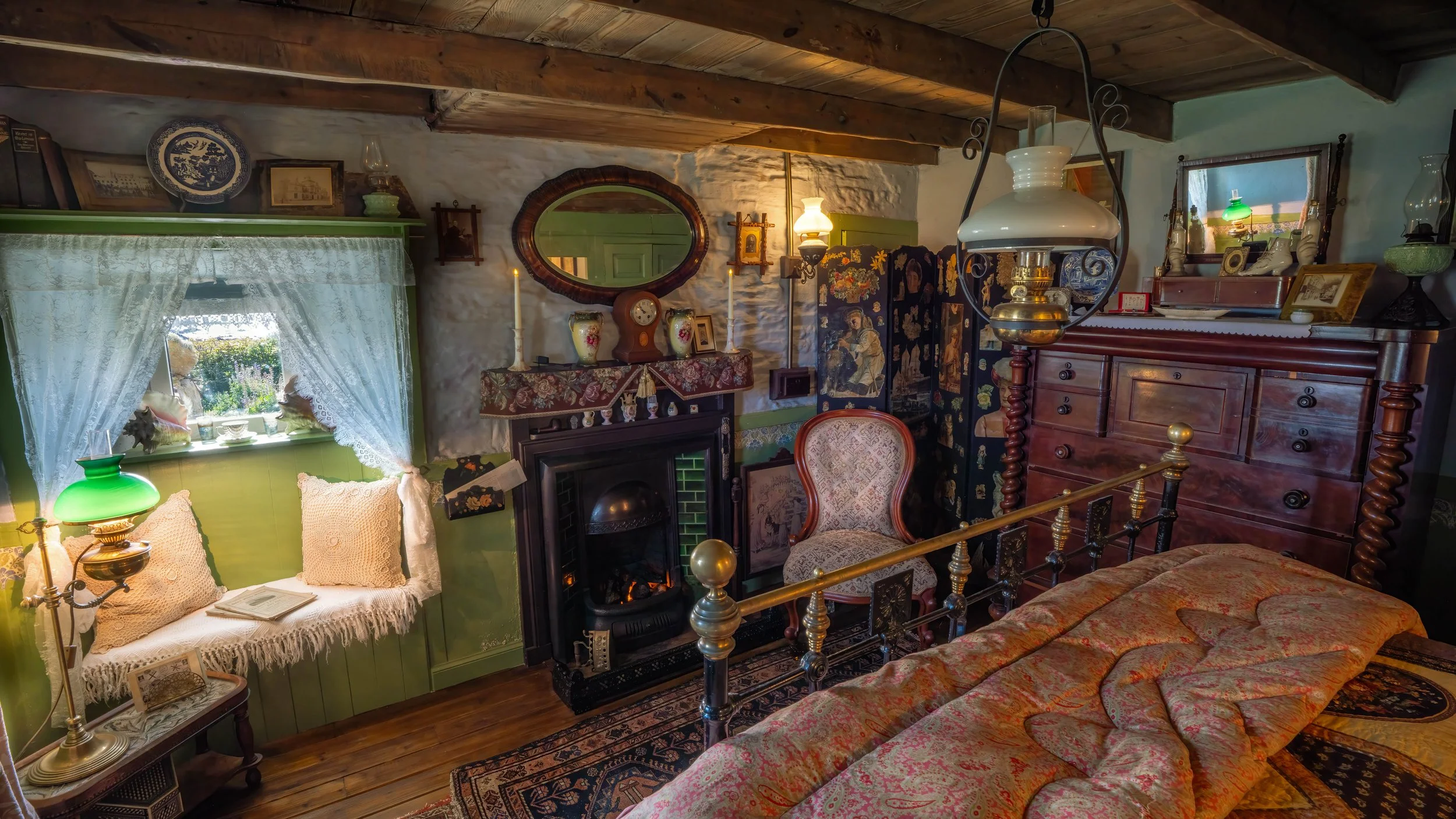 Vintage cozy bedroom with wooden ceiling beams, a window with lace curtains, a green painted bench with pillows and an open book, a fireplace with decorative vases, a floral shelf cloth, an ornate armchair, a large wooden dresser, and various antique