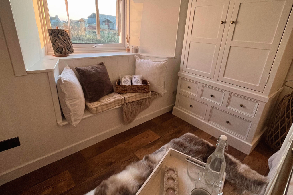 A cozy window nook with pillows and a basket of rolled towels, next to a large white cabinet, with a fur throw on a table and a glass bottle with glasses nearby.