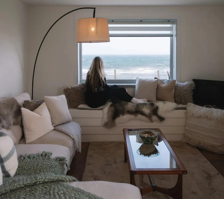 A woman with long blonde hair sitting on a window seat, gazing outside at a beach and ocean view, surrounded by various cushions and throws in a cozy living room.