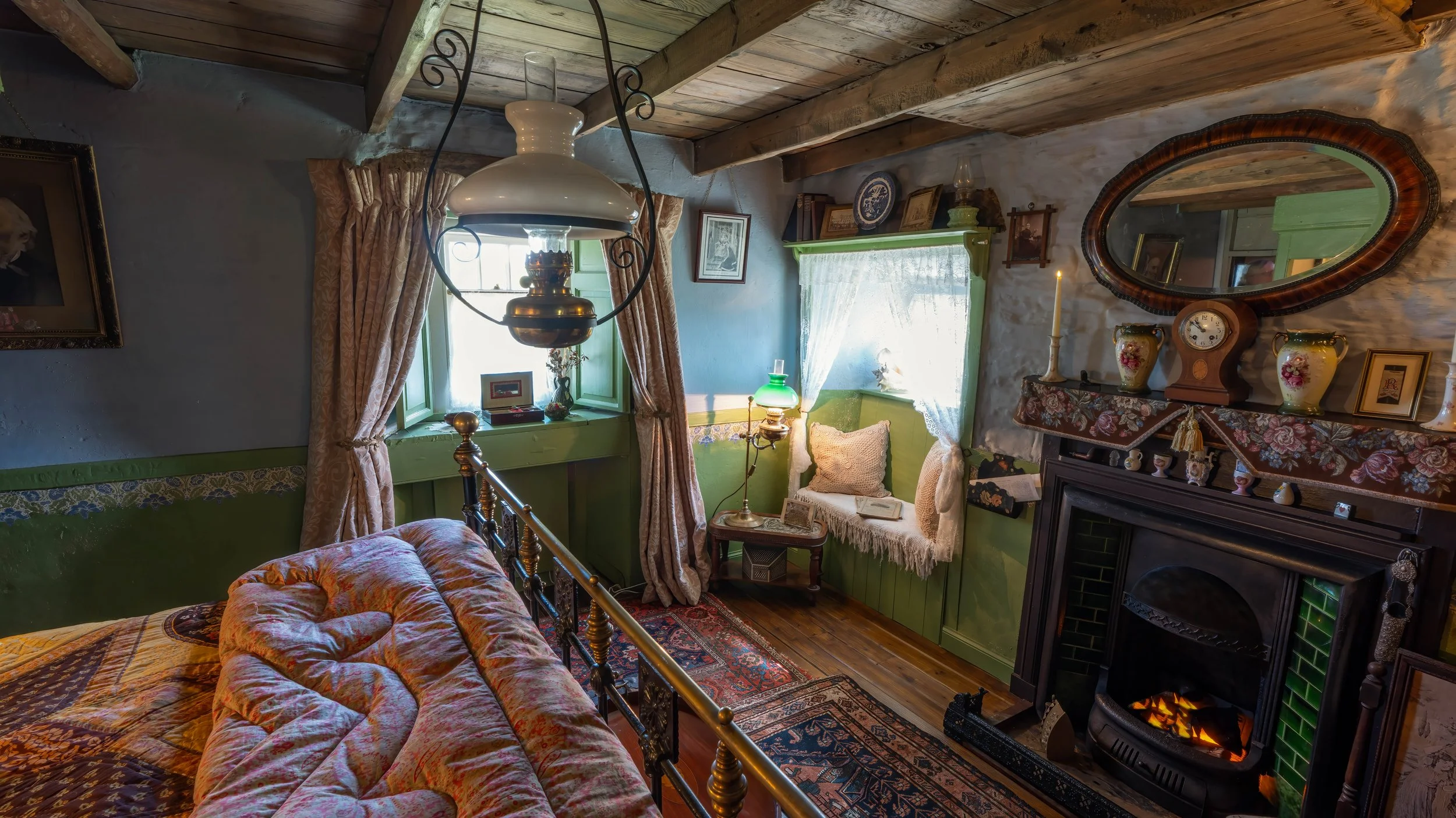 Cozy vintage bedroom in Boloquoy Victorian Farm & Watermill with wooden ceiling beams, an ornate brass bed with a floral quilt, a fireplace with decorative items, two windows with lace curtains, and antique furnishings and decor.