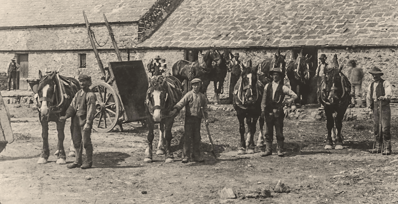Original photo of How Farm. Historical black and white photo of farmers with horses and plows in front of a stone building.