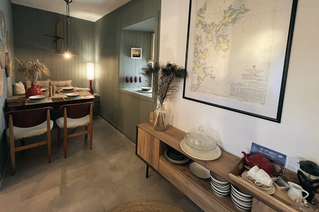 A cozy dining area with a dining table set for four, decorated with a large red vase filled with dried pampas grass. The walls are painted in green shades, with wall-mounted light fixtures, artwork, and small decorative items. A wooden sideboard hold