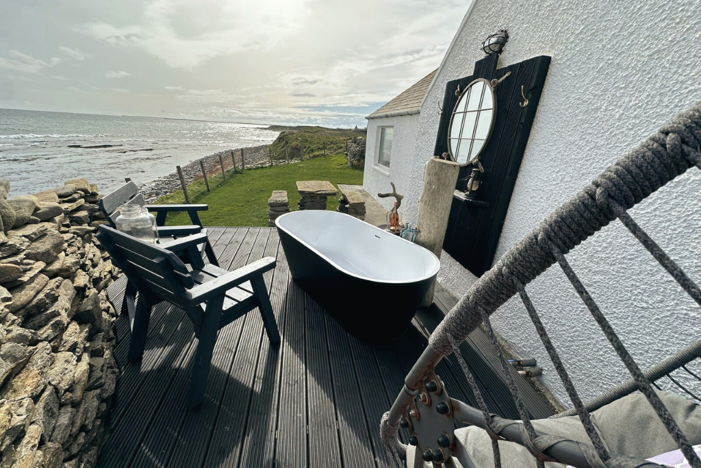 Outdoor bathroom with a freestanding bathtub, black chairs, and scenic ocean view, part of a house with white exterior walls and decorative mirrors.