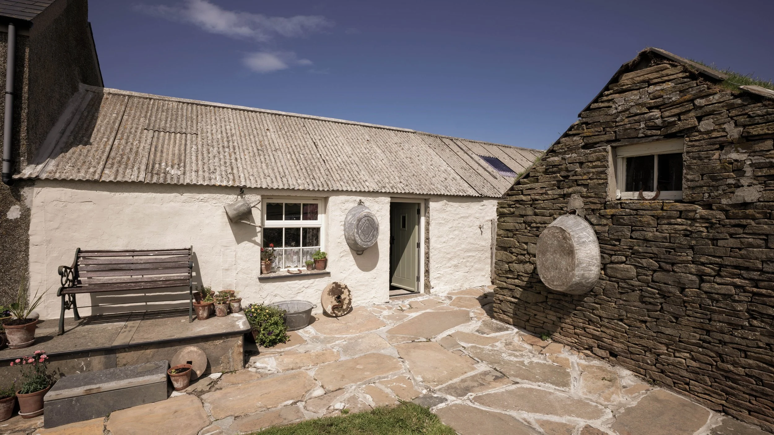 A rustic courtyard at Boloquoy Victorian Farm & Watermill. The courtyard has stone paving, potted plants, a bench, and hanging decorative pieces, under a clear blue sky.