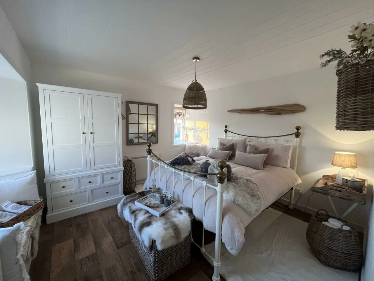 A cozy bedroom with a white iron bed frame, multiple pillows, and beige bedding. There is a nightstand with a lamp, a white wardrobe, a window with a view outside, and decorative baskets and wall art in a rustic style.