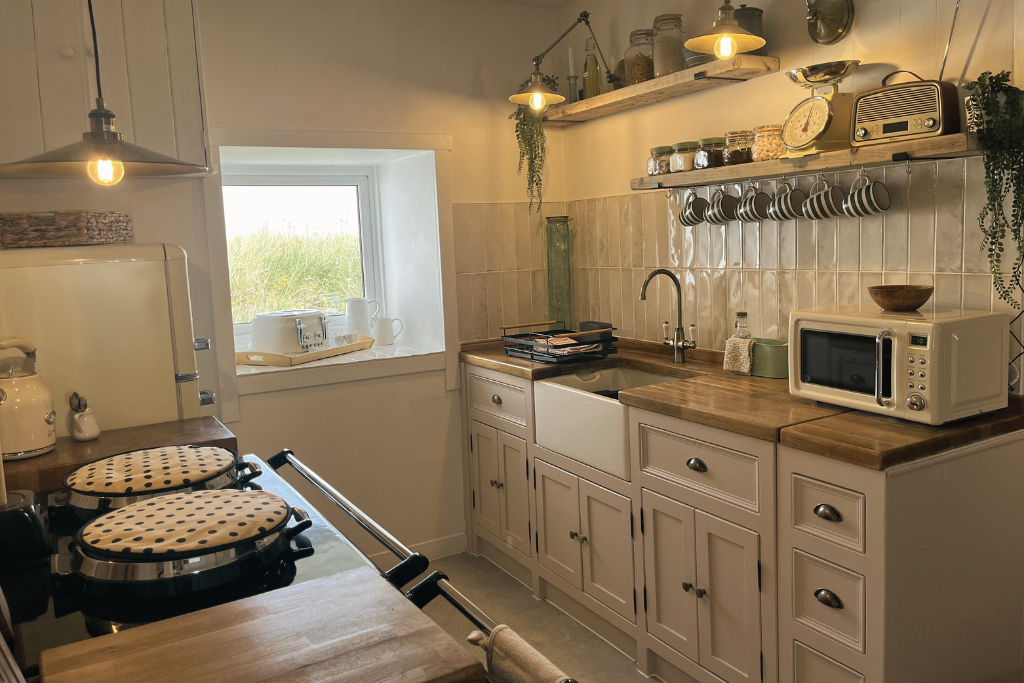 Cozy kitchen with white cabinets, wooden countertops, and a window with a view of greenery. Contains small appliances like a microwave and toaster, jars, a vintage radio, hanging cups, and decorative plants.