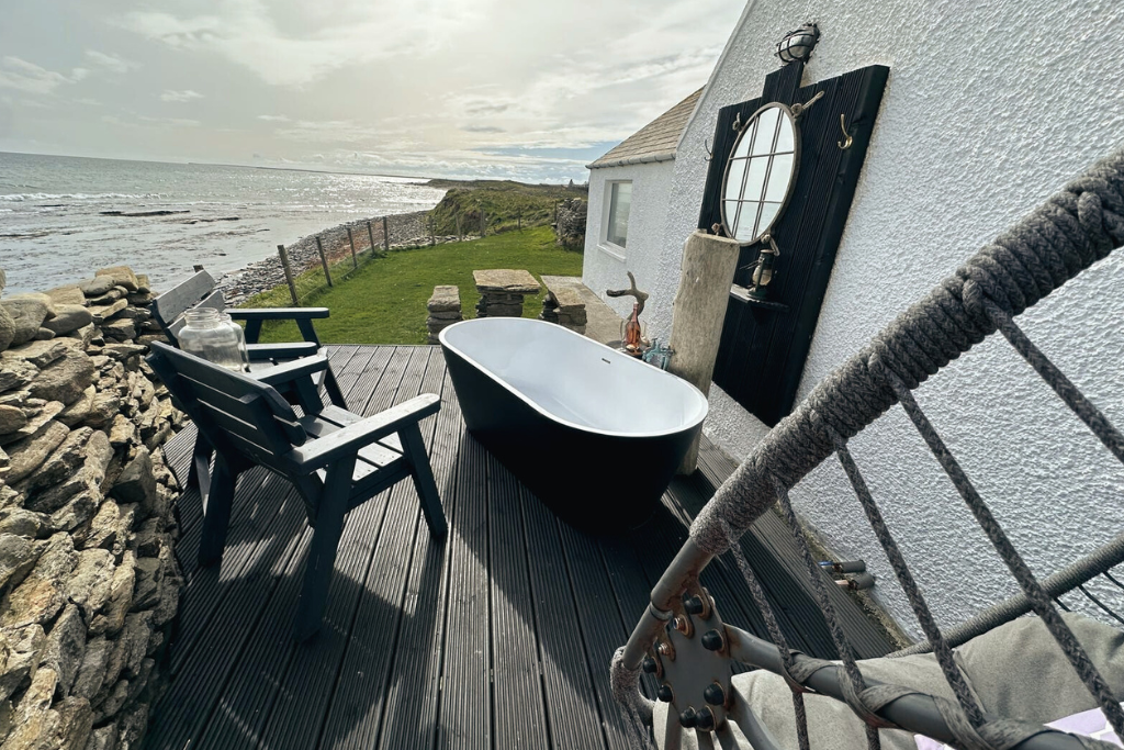Outdoor patio at Kilnbarn Cottage, with a black bathtub, two black chairs, a glass jar, and seaside view with grass, rocks, and ocean waves.
