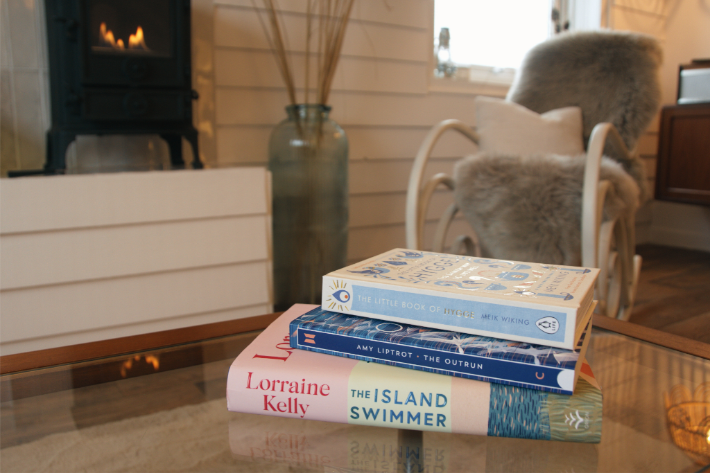 Stack of three books on a glass coffee table in a cozy living room. The top book is light blue titled 'The Little Book of Hygge,' the middle book is dark blue titled 'The Outrun,' and the bottom book is pink and white titled 'The Island Swimmer' by L