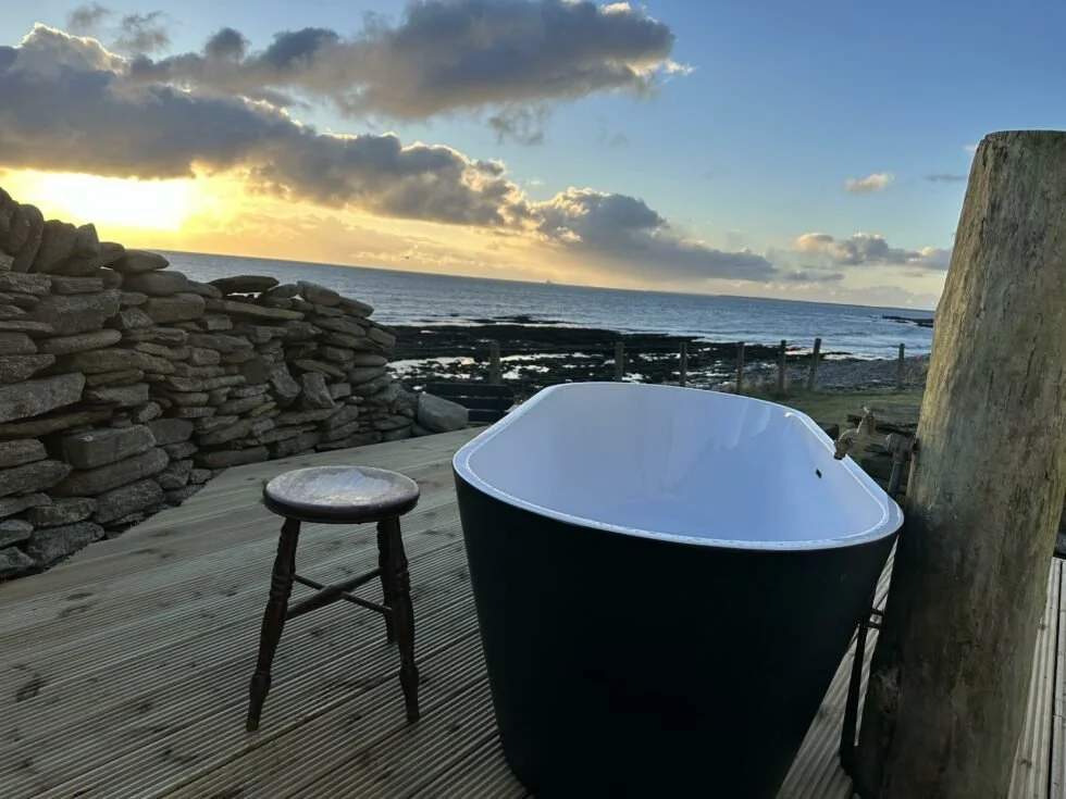 A black bathtub on a wooden deck overlooking the ocean during sunset, with a stone wall and a small wooden stool.