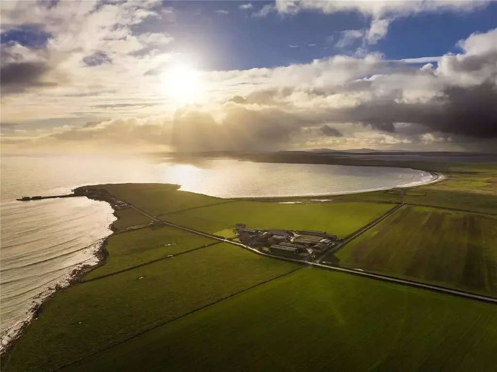 Aerial view of Sanday coastal landscape with a curved shoreline, green fields, and a few buildings under a partly cloudy sky with sunlight breaking through.