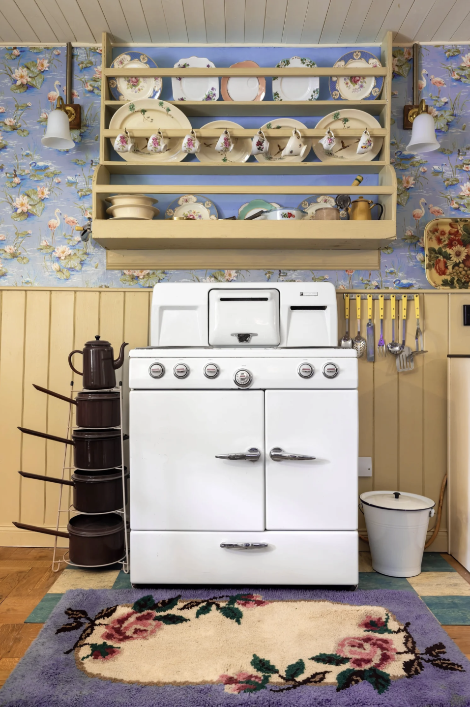 Vintage white kitchen stove in with black teapot on the left and a floral rug on the floor, yellow wooden wall paneling, shelves with decorated plates and teacups, utensils hanging on yellow hooks, floral wallpaper with flamingos and greenery.