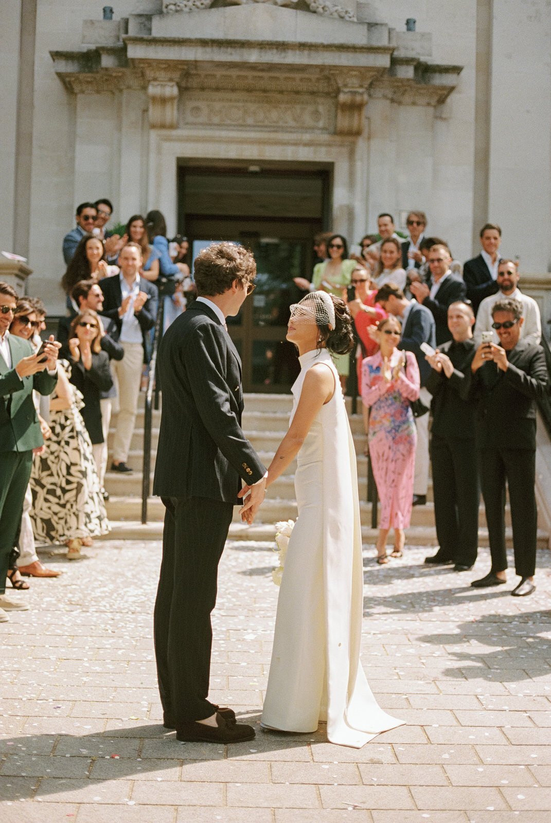 Ein Brautpaar hält Händchen bei ihrer Hochzeit im Freien vor einer Gruppe von Gästen, die sie umringen und feiern.