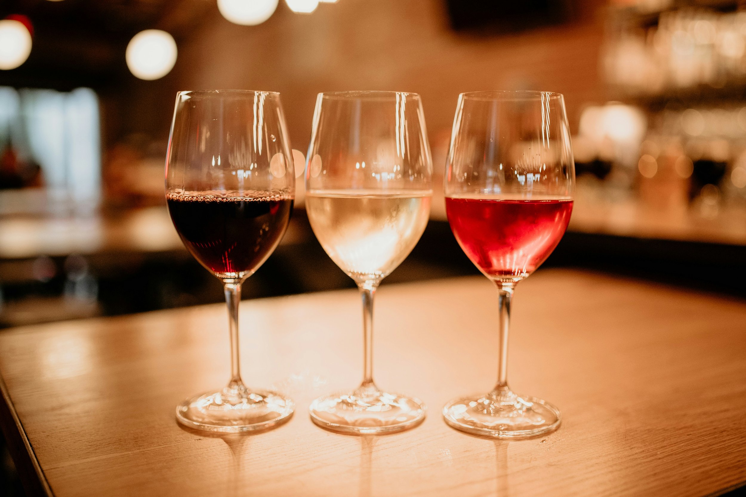 Three wine glasses on a wooden surface, filled with red wine, white wine, and rosé wine, in a dimly lit bar or restaurant setting.