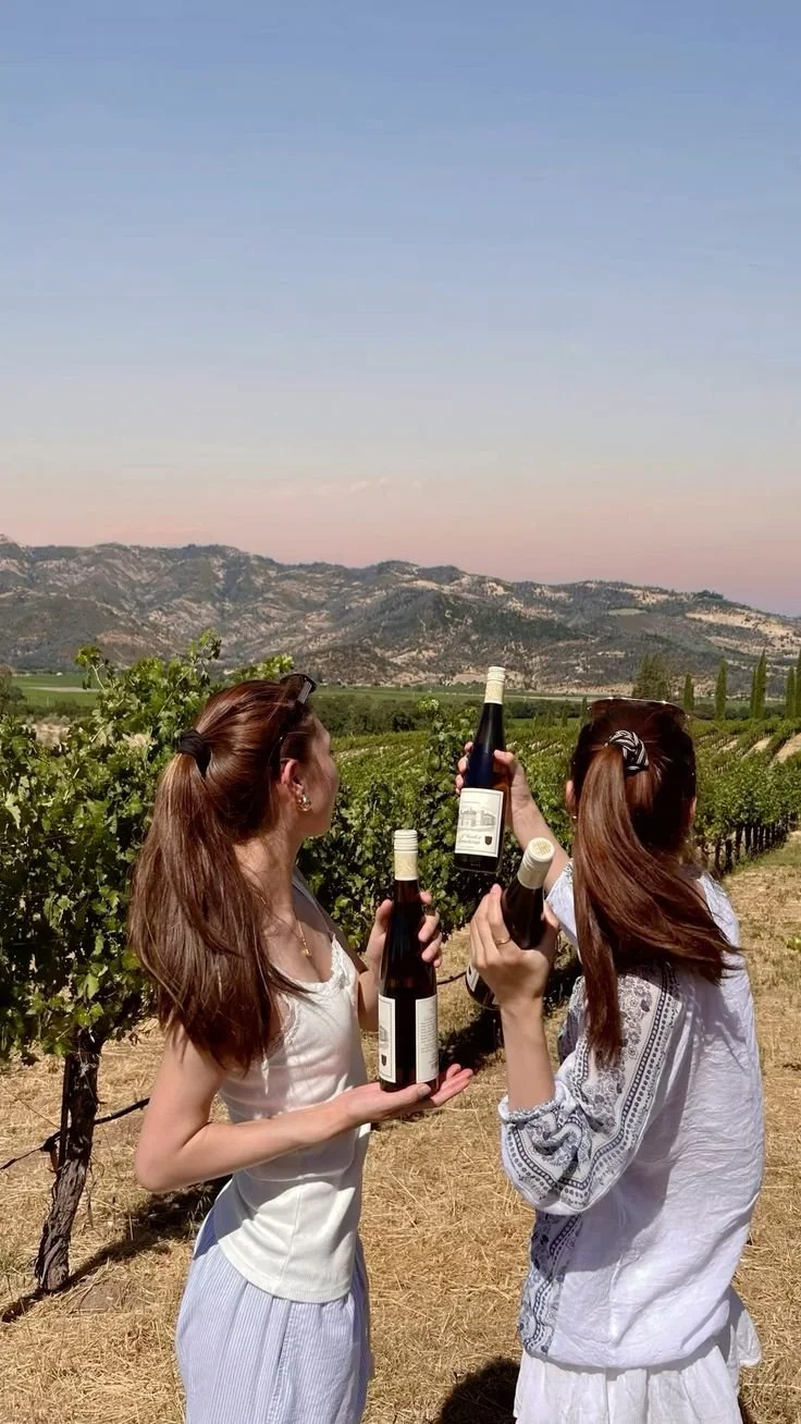 Two women in a vineyard holding up bottles of wine and celebrating against a backdrop of mountains and vineyards.