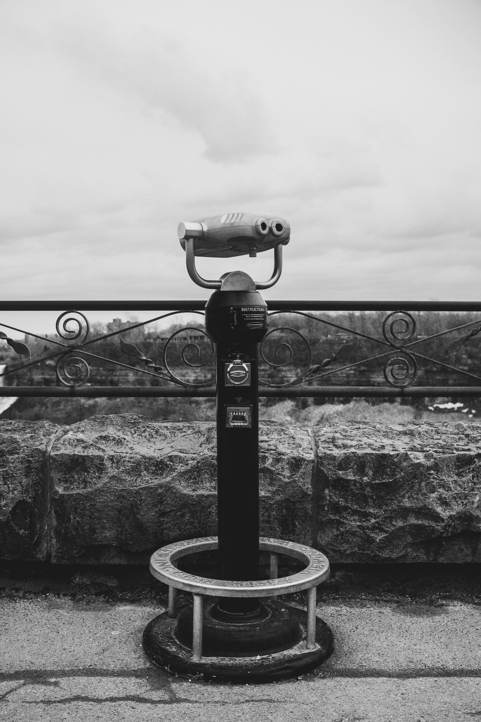 Black and white photo of a coin-operated binocular viewer on a stone platform overlooking a landscape with trees and cloudy sky.