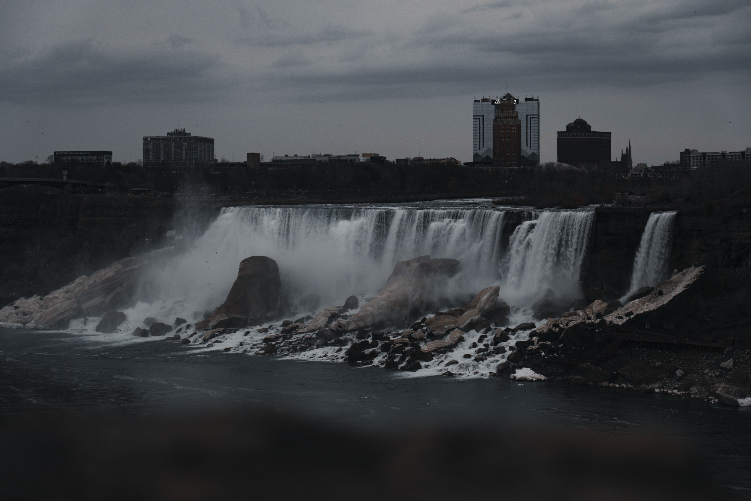 Dark, cloudy sky over waterfalls with a city skyline in the background, showing buildings and skyscrapers.