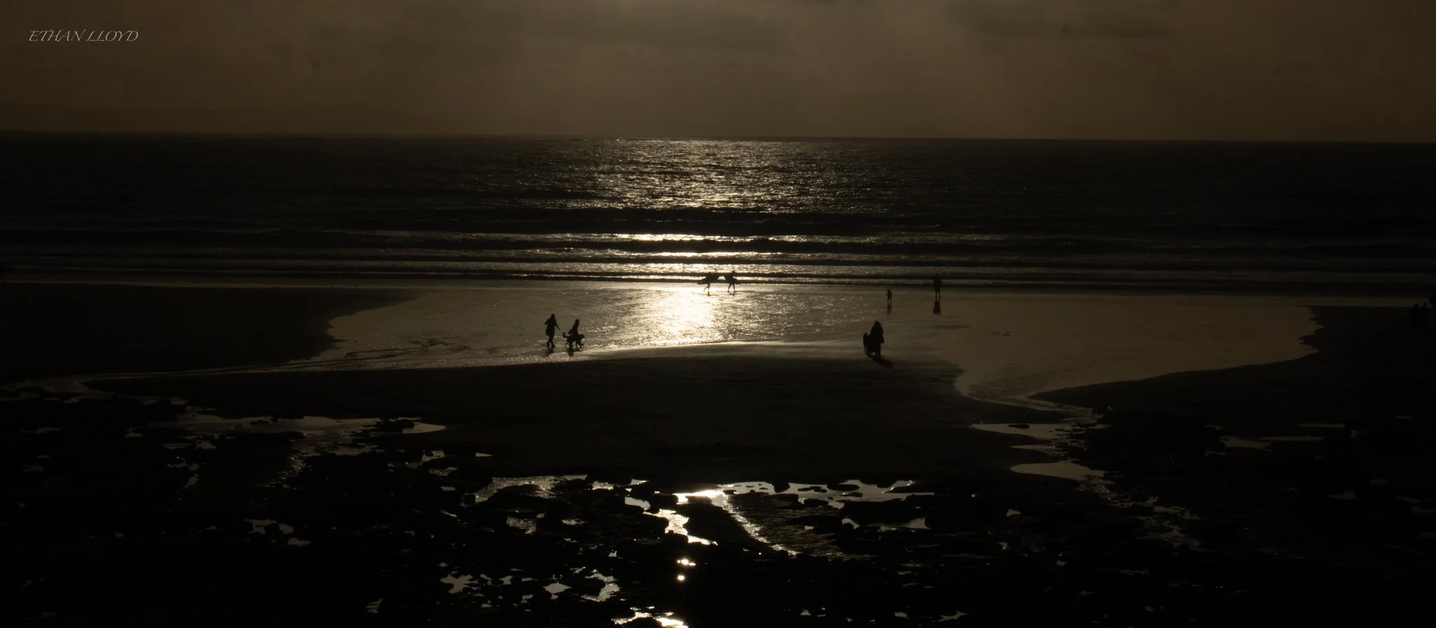 Silhouette of people walking along a beach at sunset, with waves in the background and a reflection of the sun on the water.