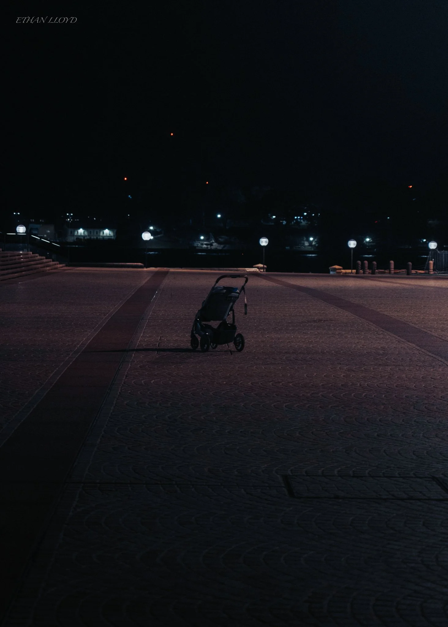 A solitary baby stroller stands on a large, empty tarmac area at night, illuminated by a few distant streetlights. The sky is dark, and no people are visible in the scene.