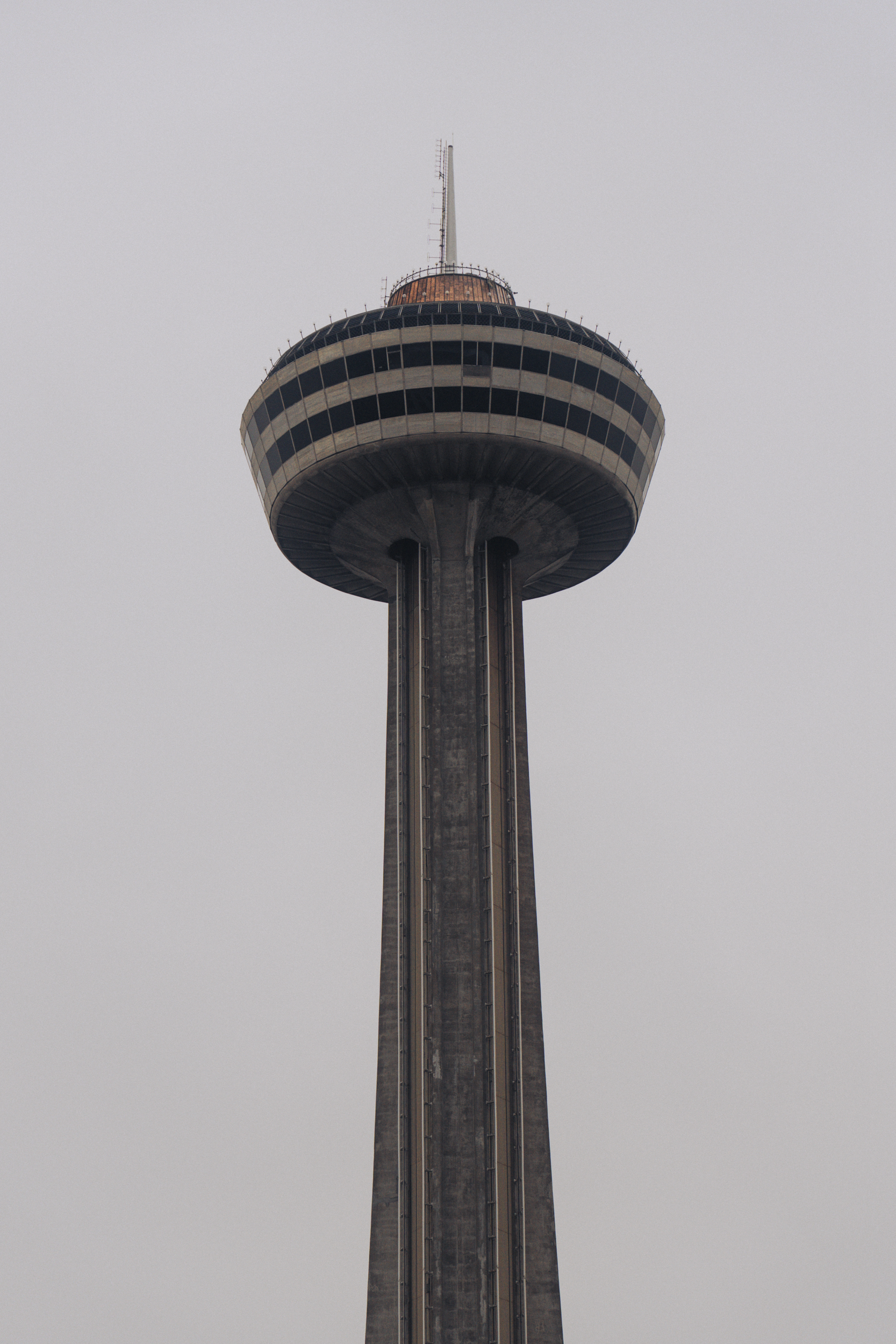 A tall observation tower with a circular viewing platform near the top, featuring a narrow base and a wider top structure, against a cloudy sky.