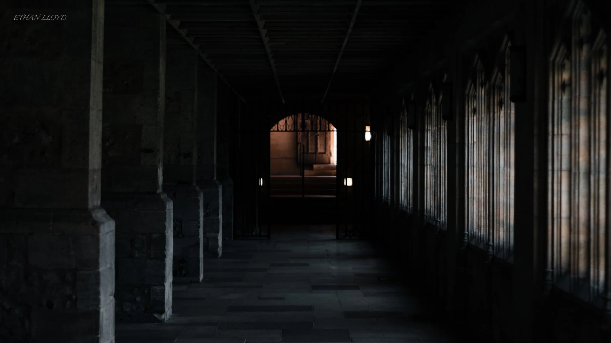 Dark corridor inside a building with stone columns on the left, windows on the right, and a door at the end leading outside where stairs are visible.