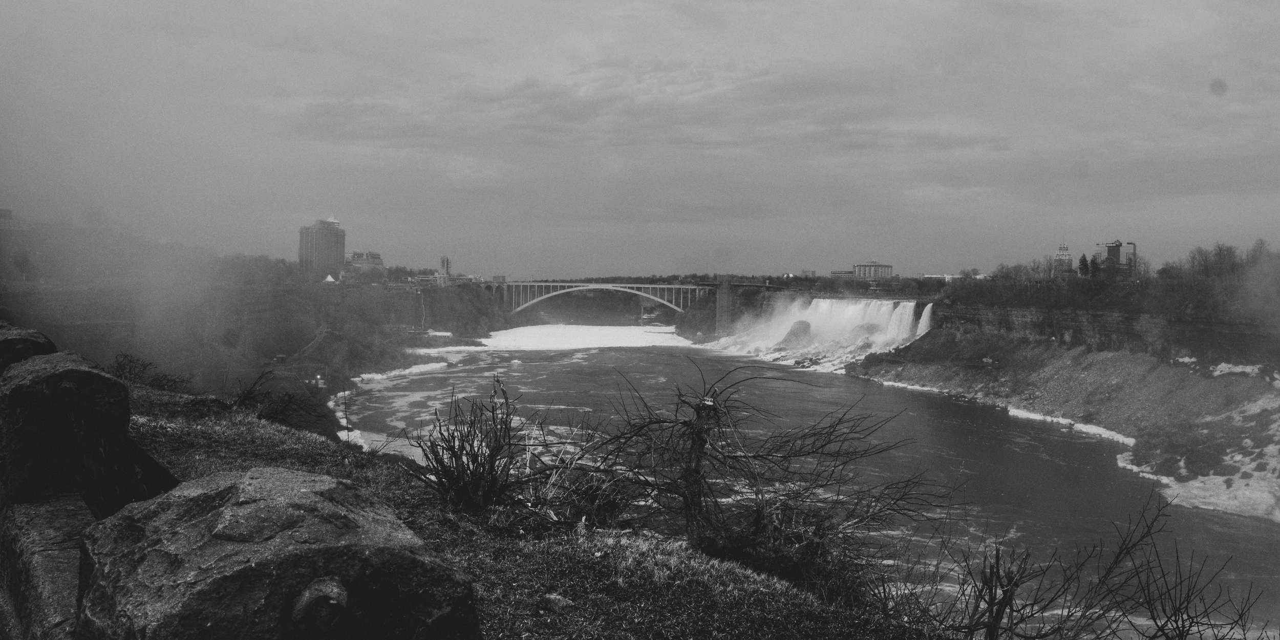 Black and white photo of Niagara Falls with the city skyline and bridge in the background, taken from the rocky shoreline.