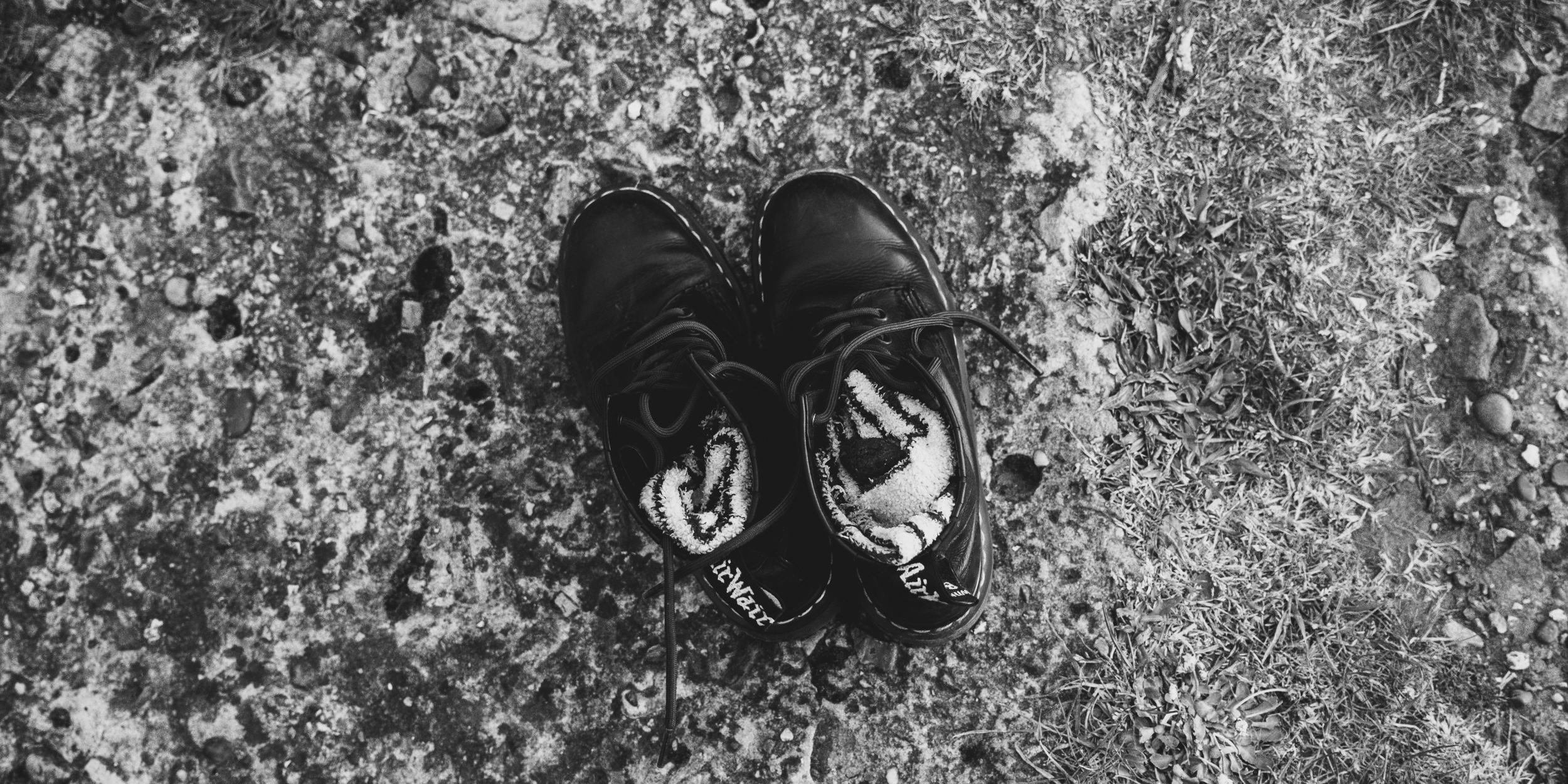 A pair of black hiking boots with white inside lining, placed on a dirt trail with some grass on the side, seen from above.