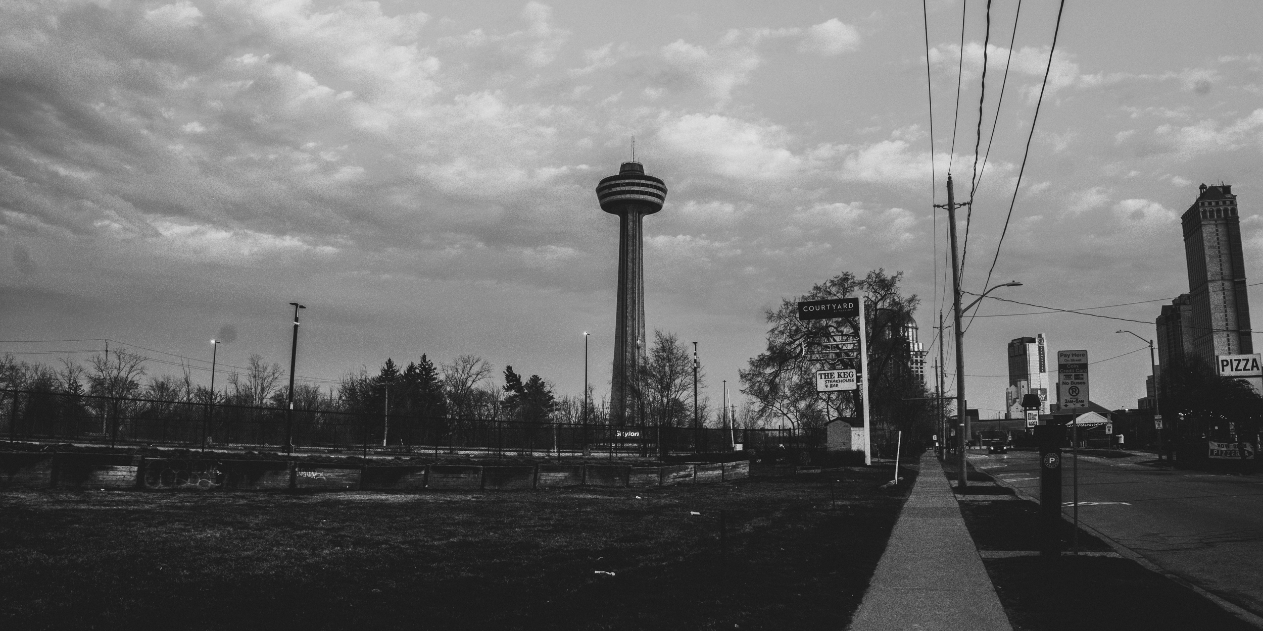 A black and white photo of an urban scene with a tall observation tower and a skyline of buildings in the background. The foreground includes a sidewalk, utility poles, and street signs.