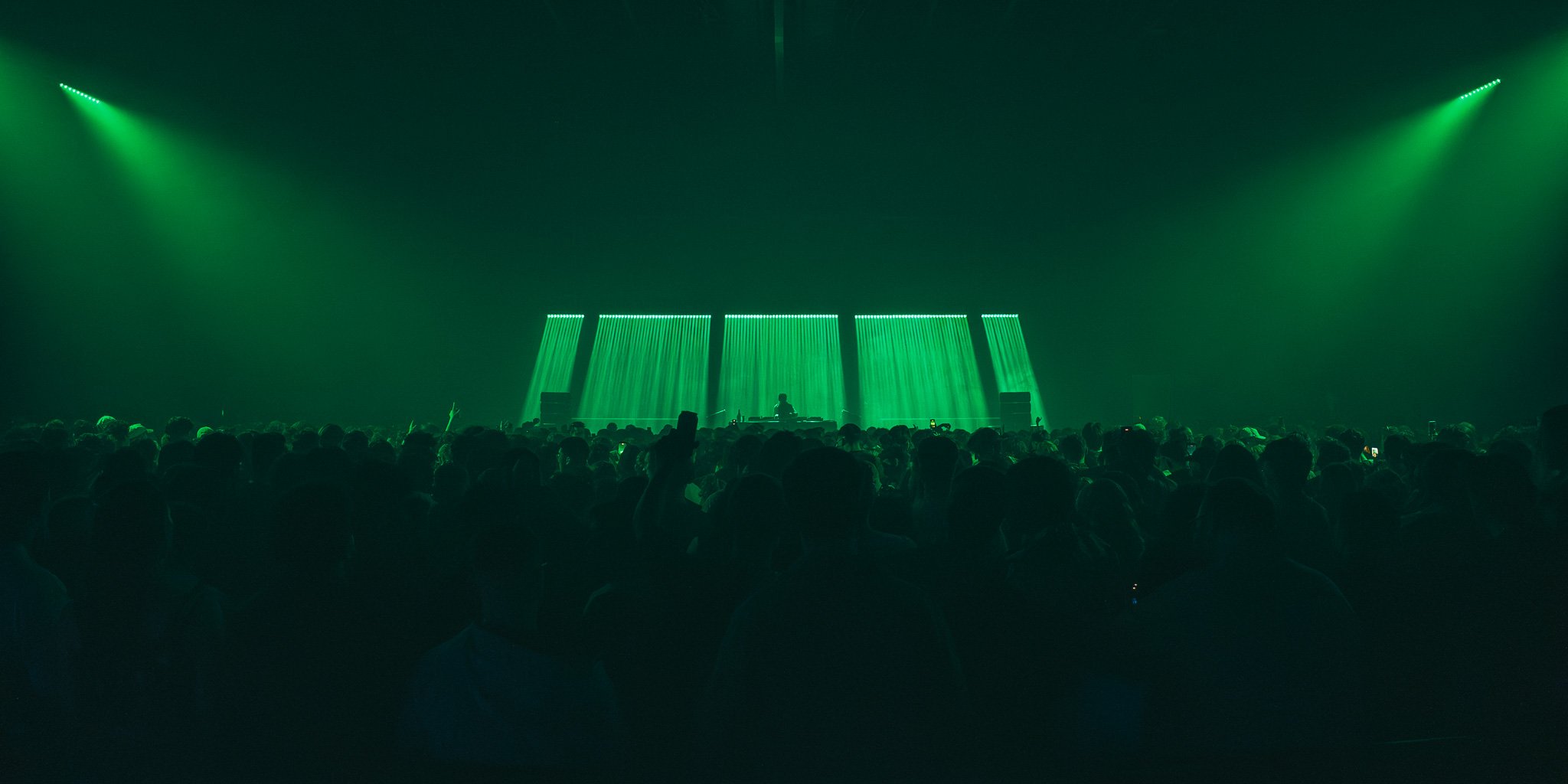 A concert scene with green lighting, a DJ or performer on stage behind a console, and a large crowd of people gathered in front.