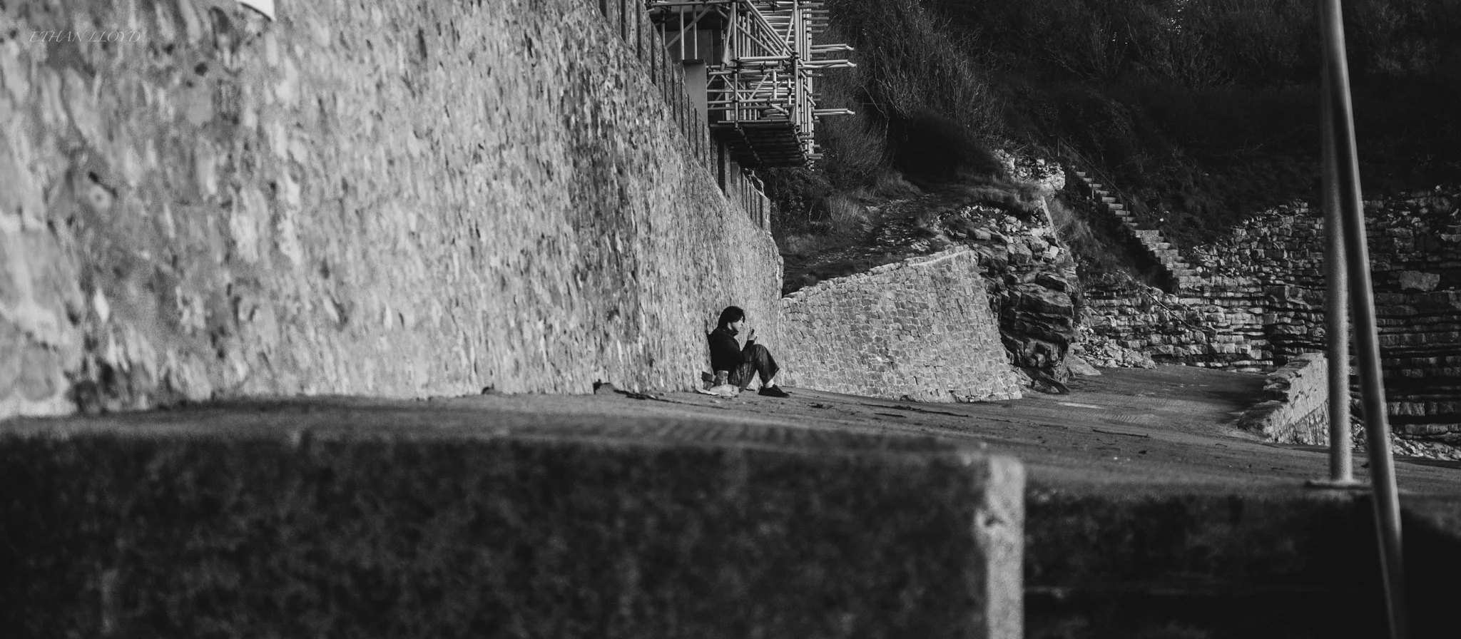 A person sitting on the ground near a stone wall, looking at a smartphone, with a pathway and stairs in the background.