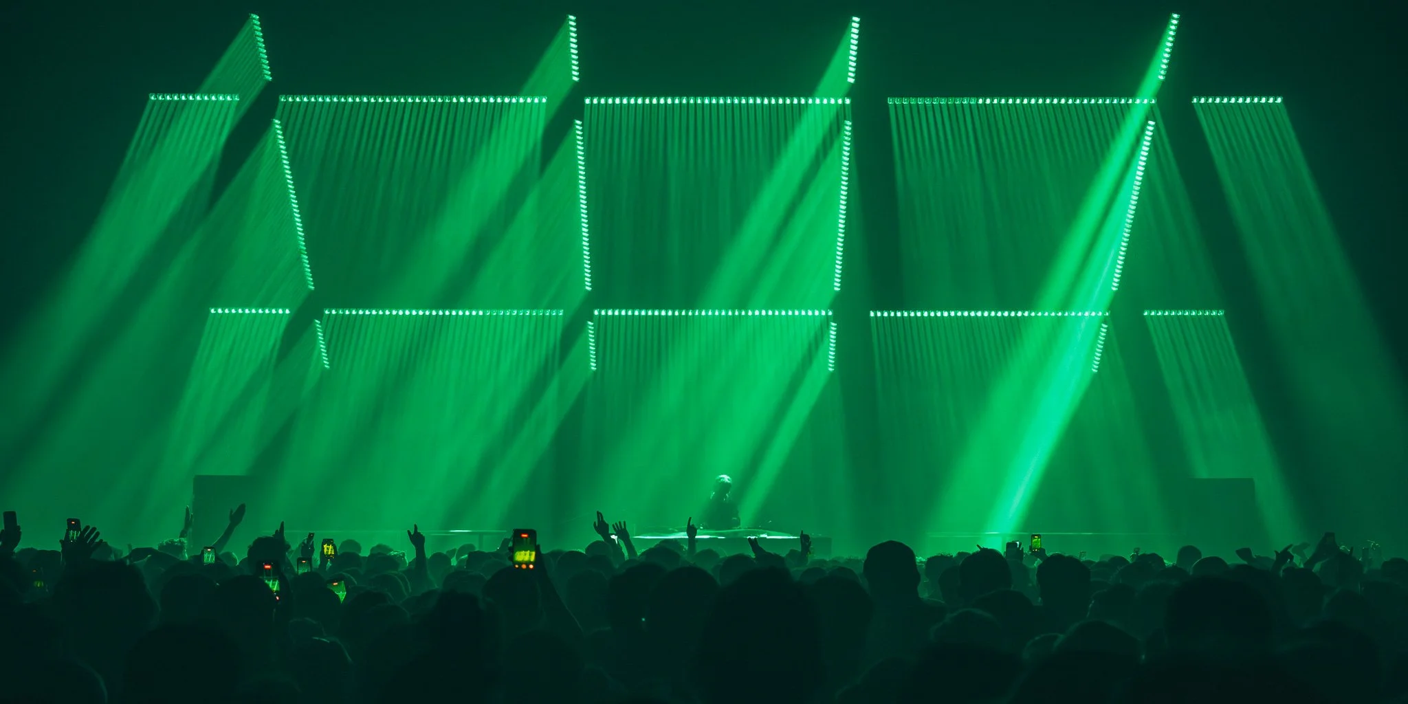 A live concert with green stage lighting, a DJ or performer in the center, and a crowded audience taking photos and videos.
