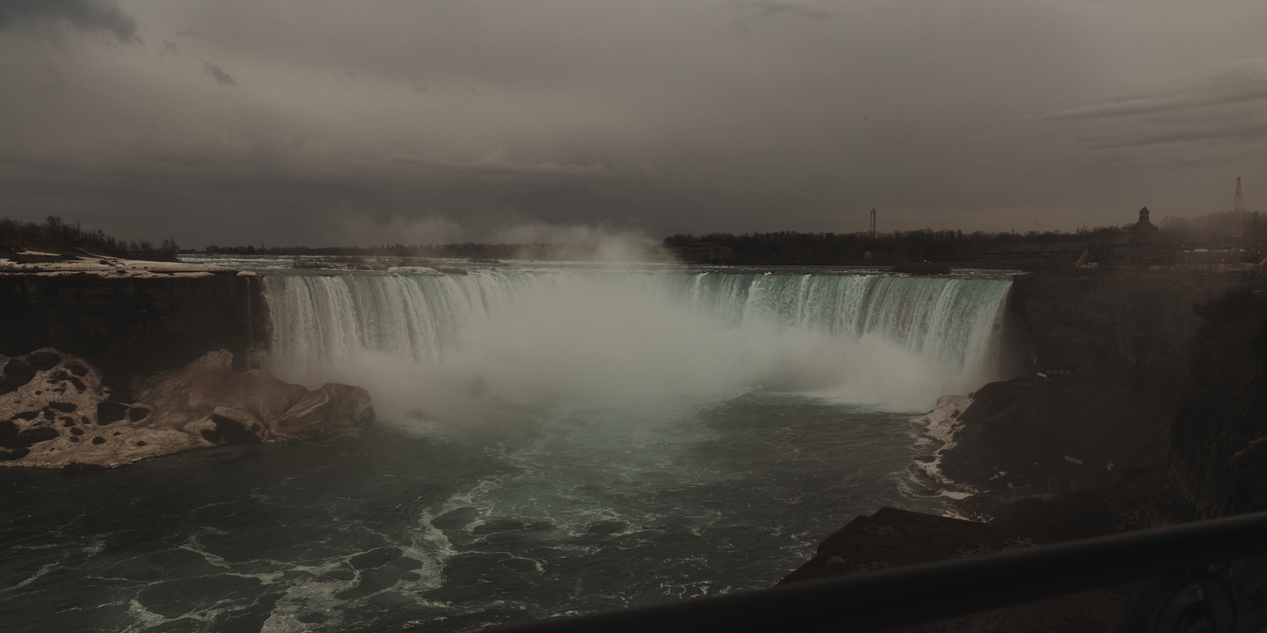 Waterfall with a wide cascade, mist rising, over a rocky riverbank under a cloudy, dark sky.