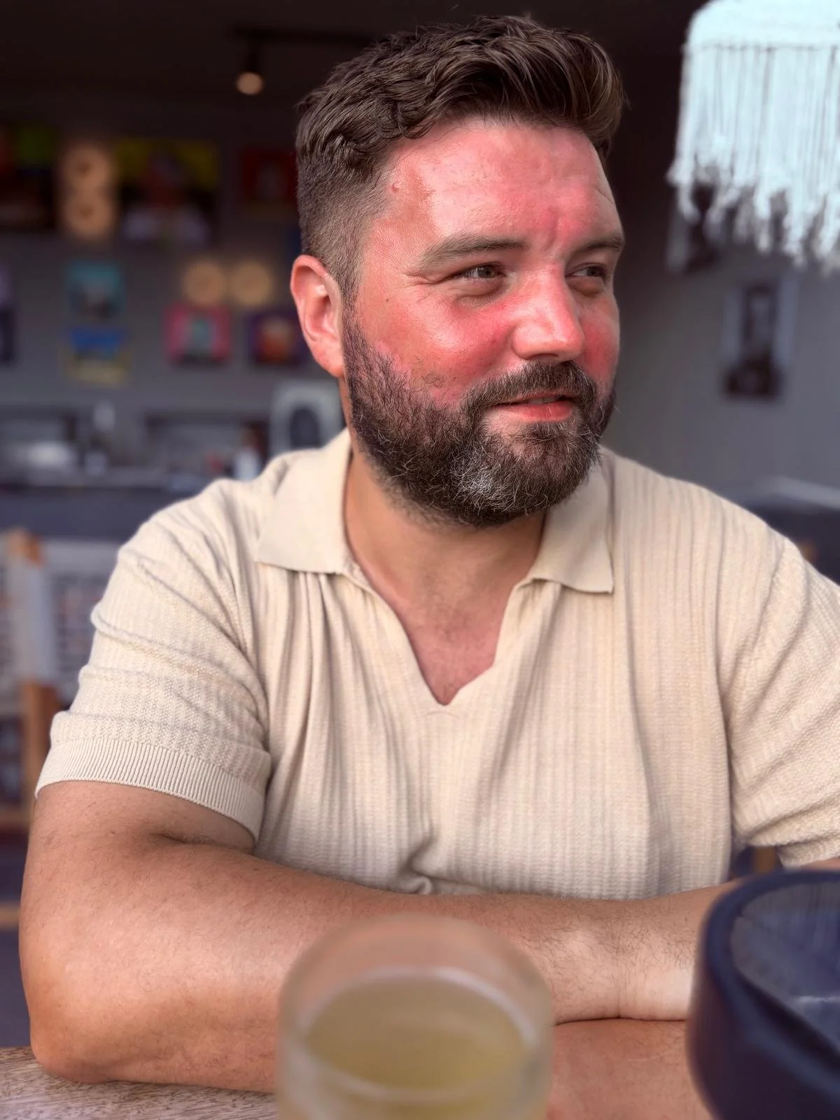 A man with a beard and short brown hair sitting at a table, looking to his right with a slight smile, in a casual indoor setting.