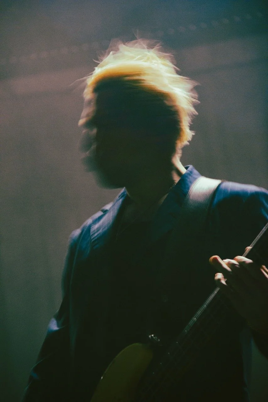 Silhouette of Queens Of The Stone Age bassist Micheal Shuman performing with the band in 2025. Image shows his energetic head movements and lighting highlighting his blonde hair waving around.