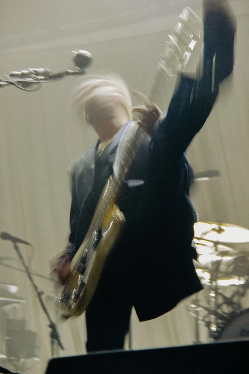 Queens Of The Stone Age bassist Micheal Shuman, in a suit, performing with the band in 2025. A purposefully blurry image capturing the energetic movements he has on stage.