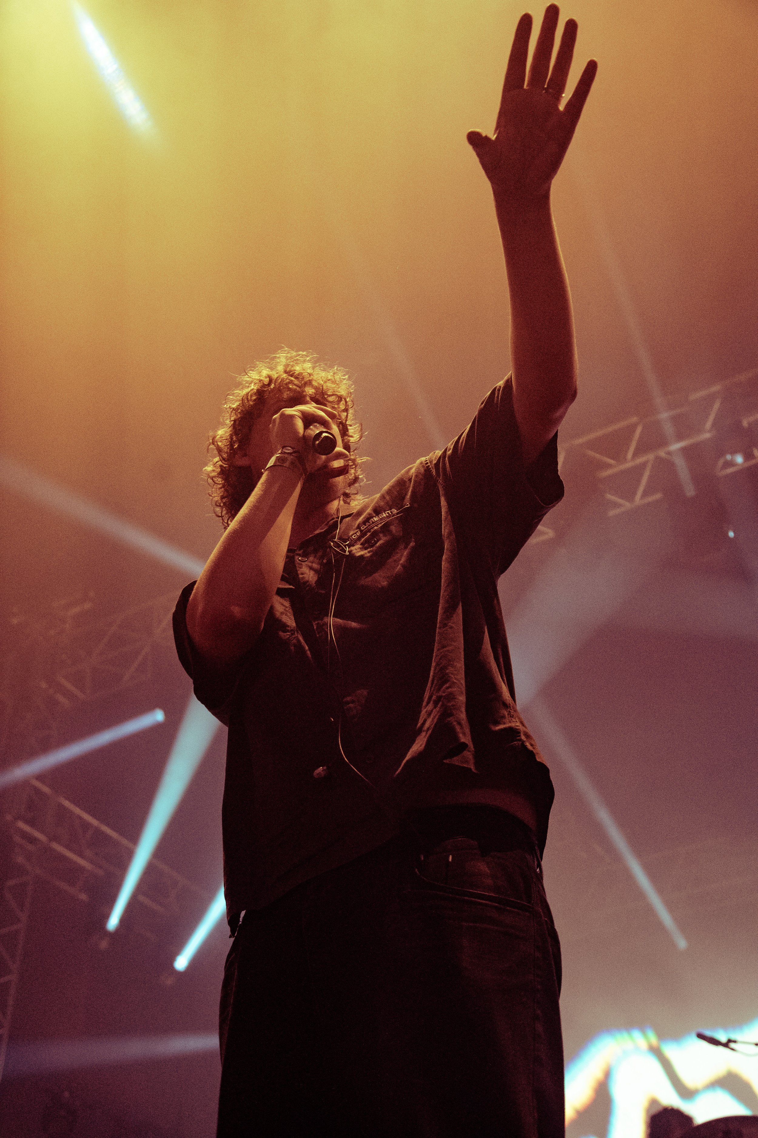 The singer of the band Fat Dog stood at the front of the stage raising his hand toward fans as the orange lights beam down on him from above.