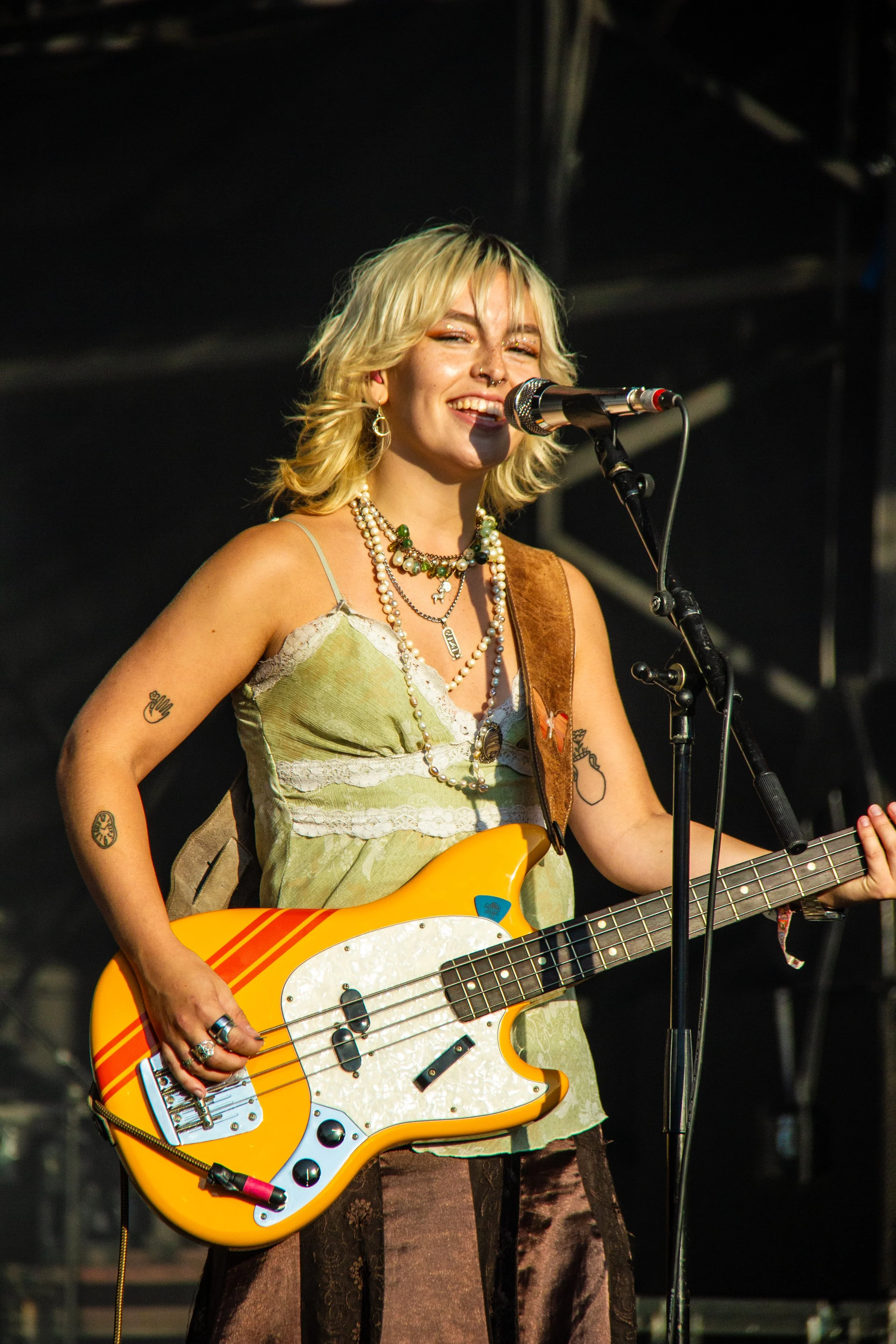 Bassist playing with the singer Nieve Ella in the bright evening sun at Camp Bestival 2025. Playing her orange Fender Vintera II 70's Competition Mustang Bass.