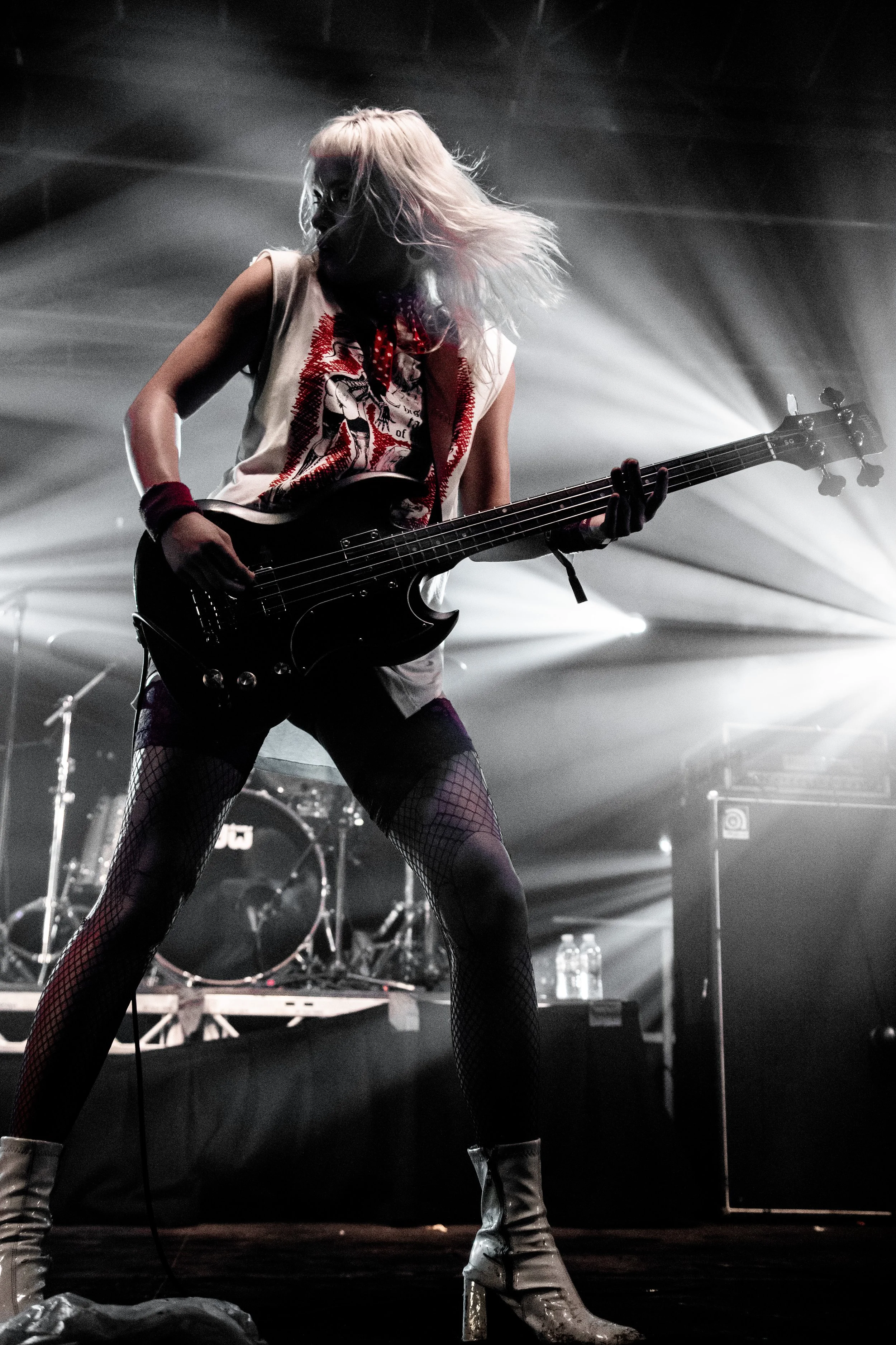 Bassist of the band The Molotovs playing at Rock and Roll Circus Festival 2026, with dramatic lighting, wearing fishnet stockings, high-heeled boots, a sleeveless t-shirt, and wristbands, with hair flowing.