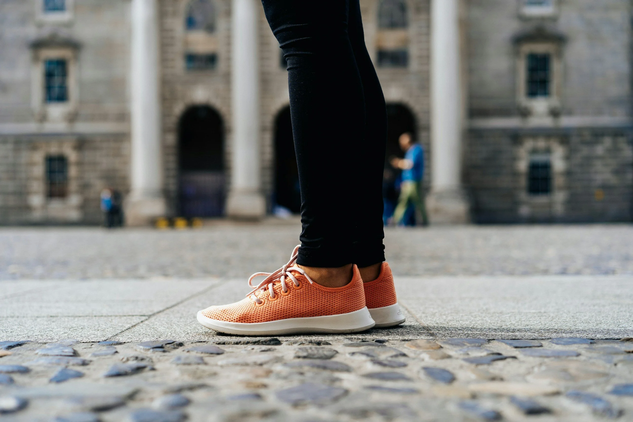 Close-up of a person wearing peach athletic shoes and black pants standing on a stone-paved sidewalk in front of a historic stone building with columns.