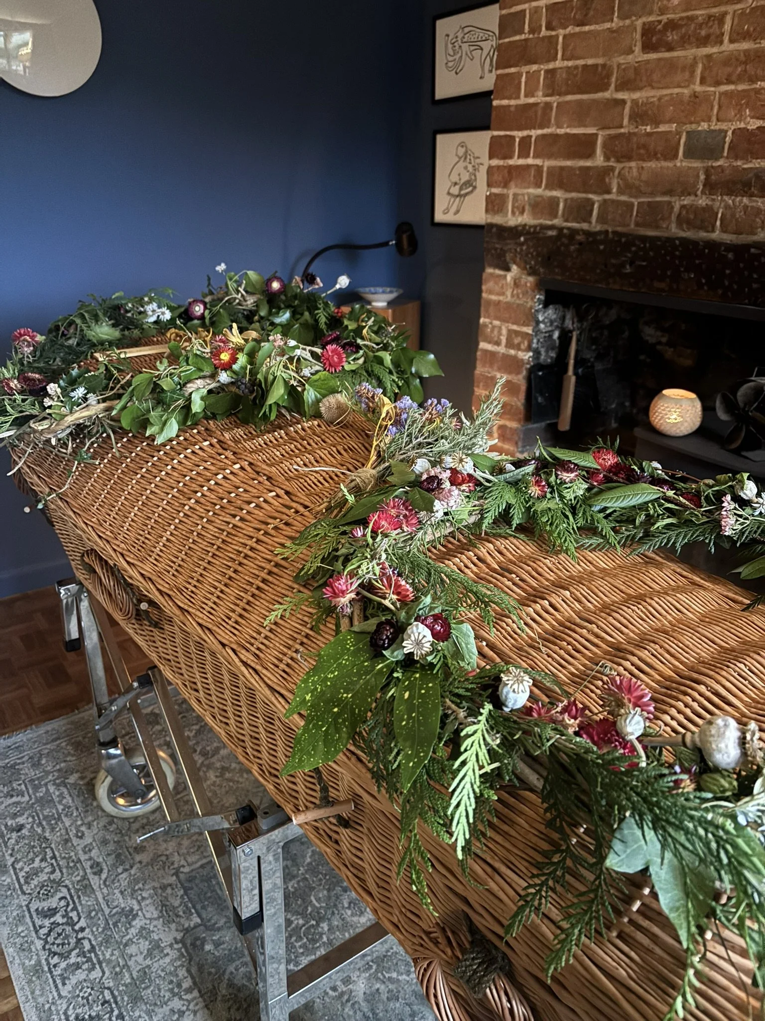 Wicker casket decorated with green foliage and colorful flowers, situated in a room with brick fireplace and blue wall.