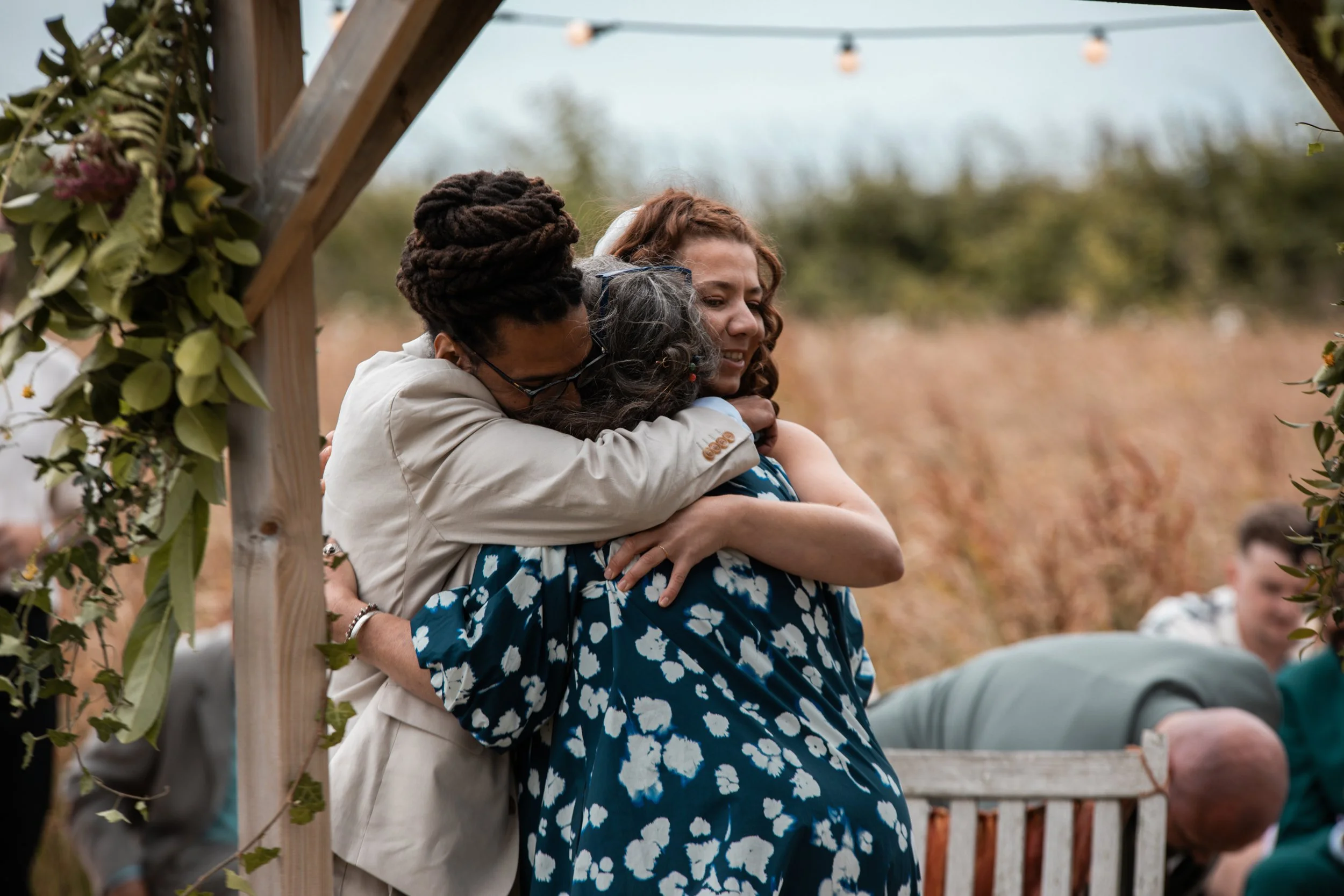 Two women hugging at an outdoor event with a wooden arch decorated with greenery and pink flowers.
