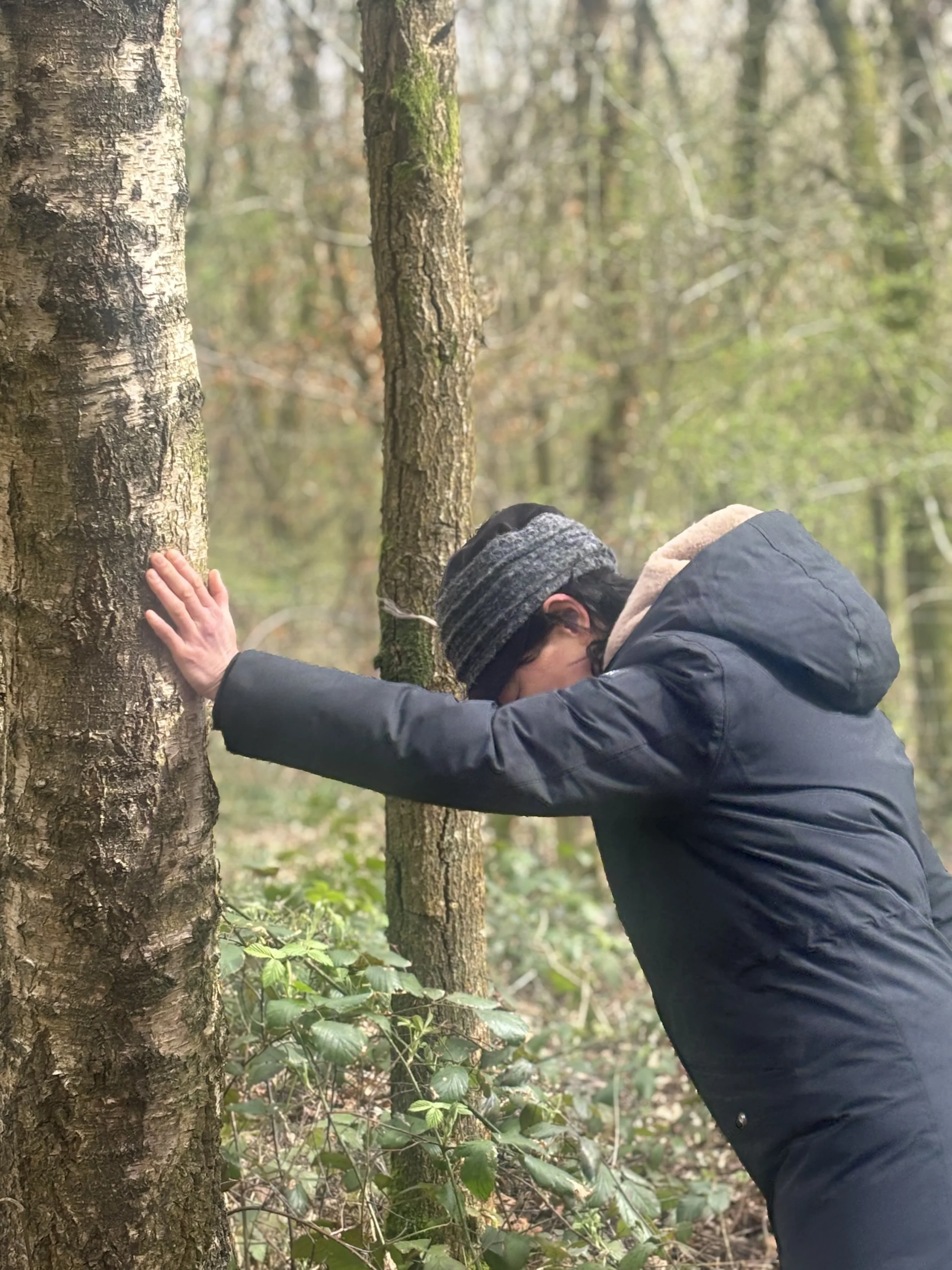 Person wearing a beanie and jacket with hood, leaning forward with forehead and hands against a tree in a forest.