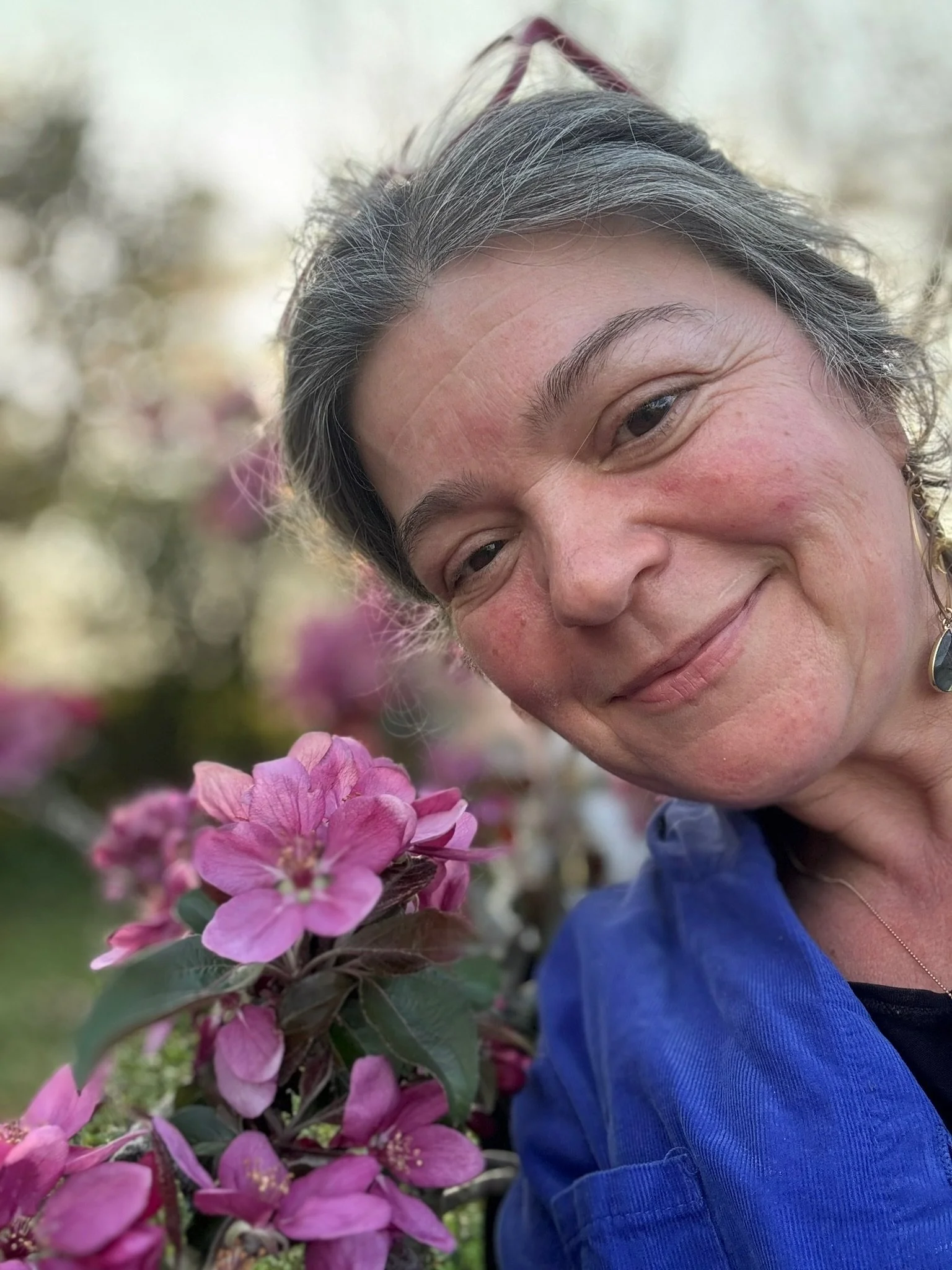 A woman with gray hair, smiling, taking a selfie outdoors near pink flowers with a blurred background.
