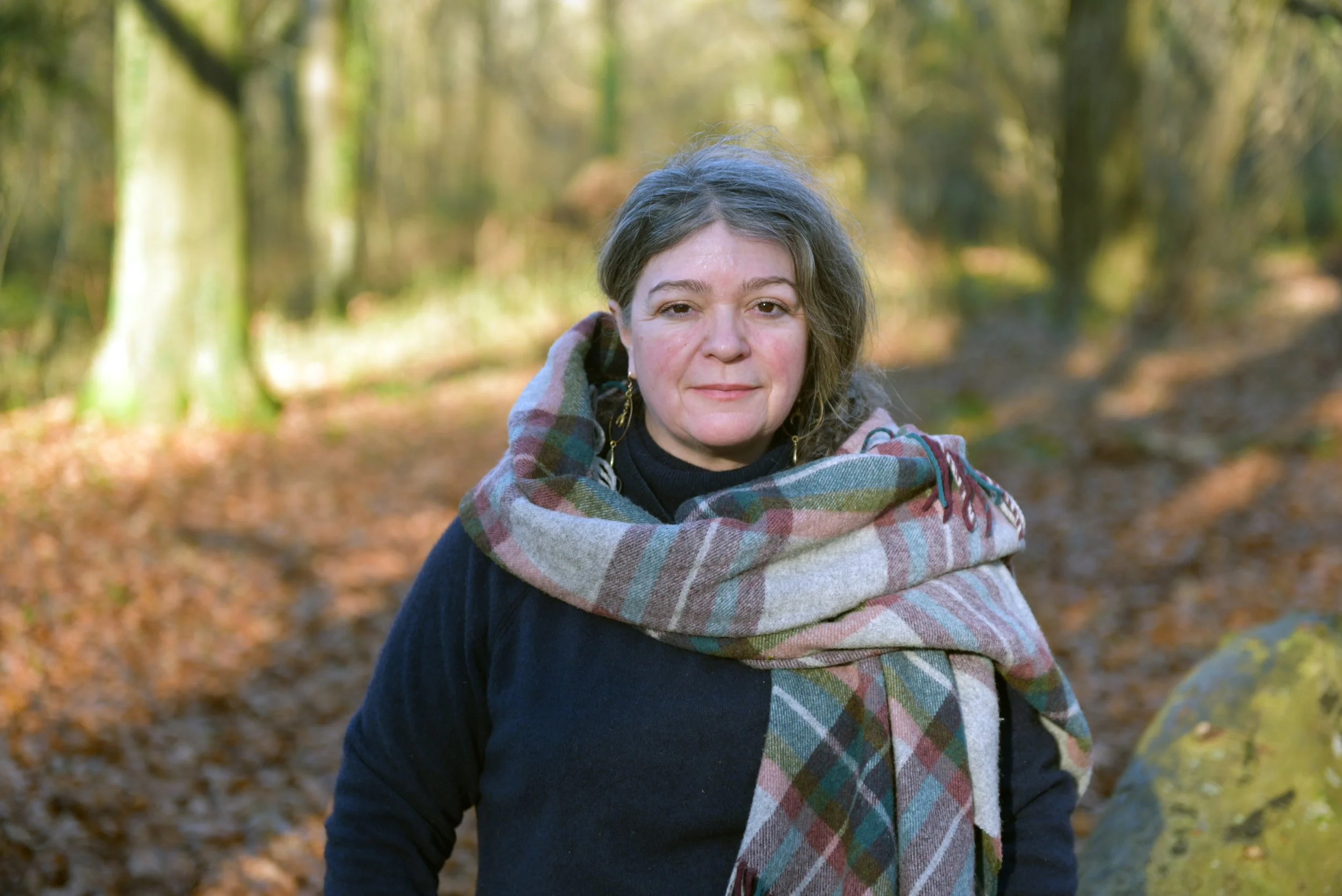A woman standing outdoors in a forest, wearing a dark top and a large plaid scarf, with trees and fallen leaves in the background.