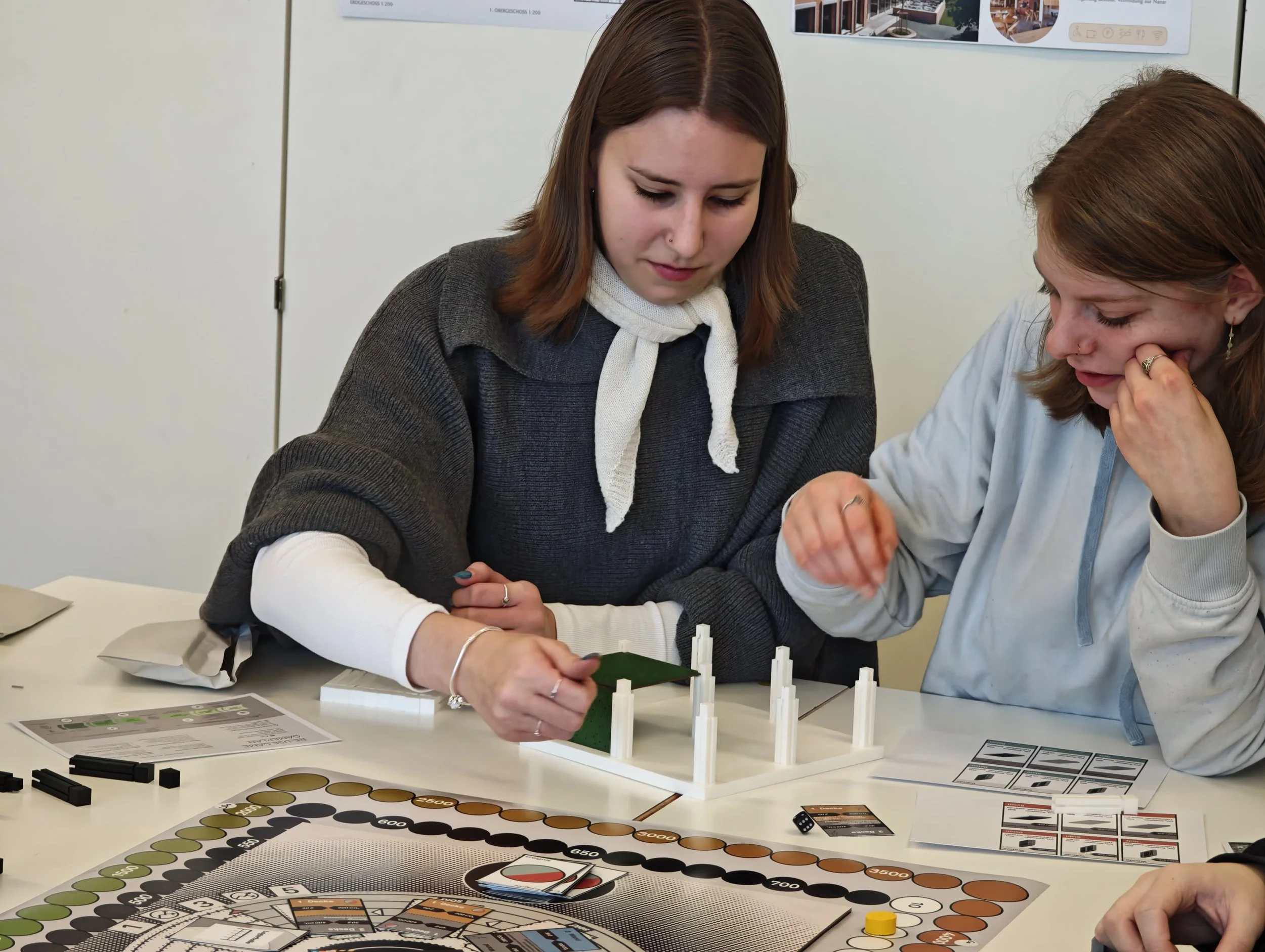 Zwei Frauen spielen ein Gesellschaftsspiel an einem Tisch mit Spielkarten, Spielsteinen und einer Spielmatte.