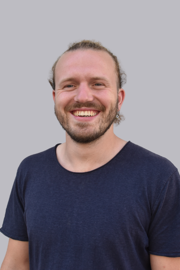 Uomo sorridente con i capelli lunghi in maglietta blu su sfondo grigio.