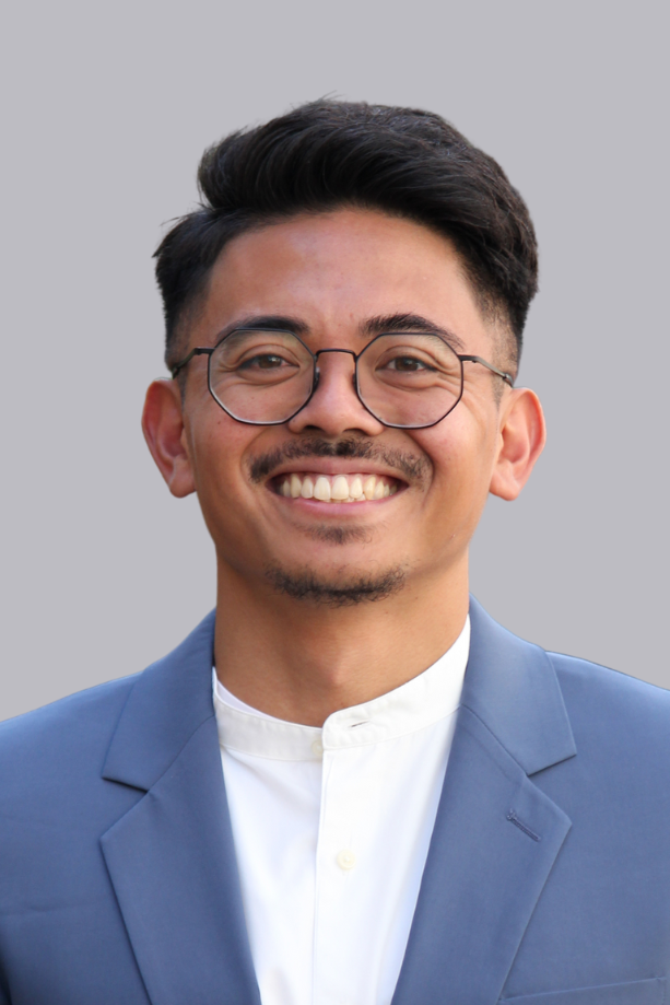 Uomo sorridente con gli occhiali, capelli scuri, in completo blu e camicia bianca, su uno sfondo grigio.