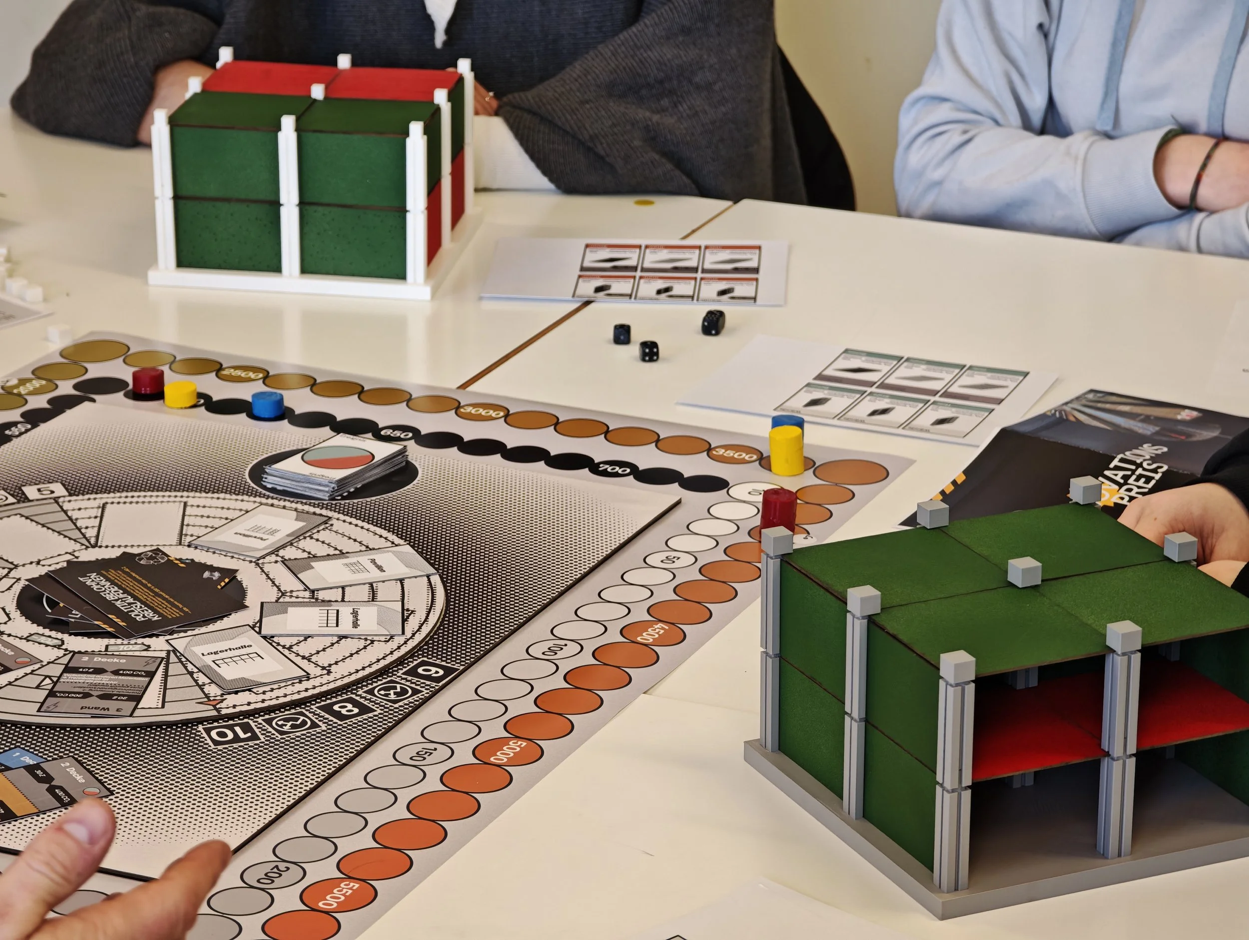 Tisch mit Brettspielen, Würfeln und Spielsteinen, darunter ein Rubik's Cube. Ein 3D-Würfelmodell und Spielkarten sind sichtbar, während eine Statue eines Gebäudes auf der rechten Seite steht.