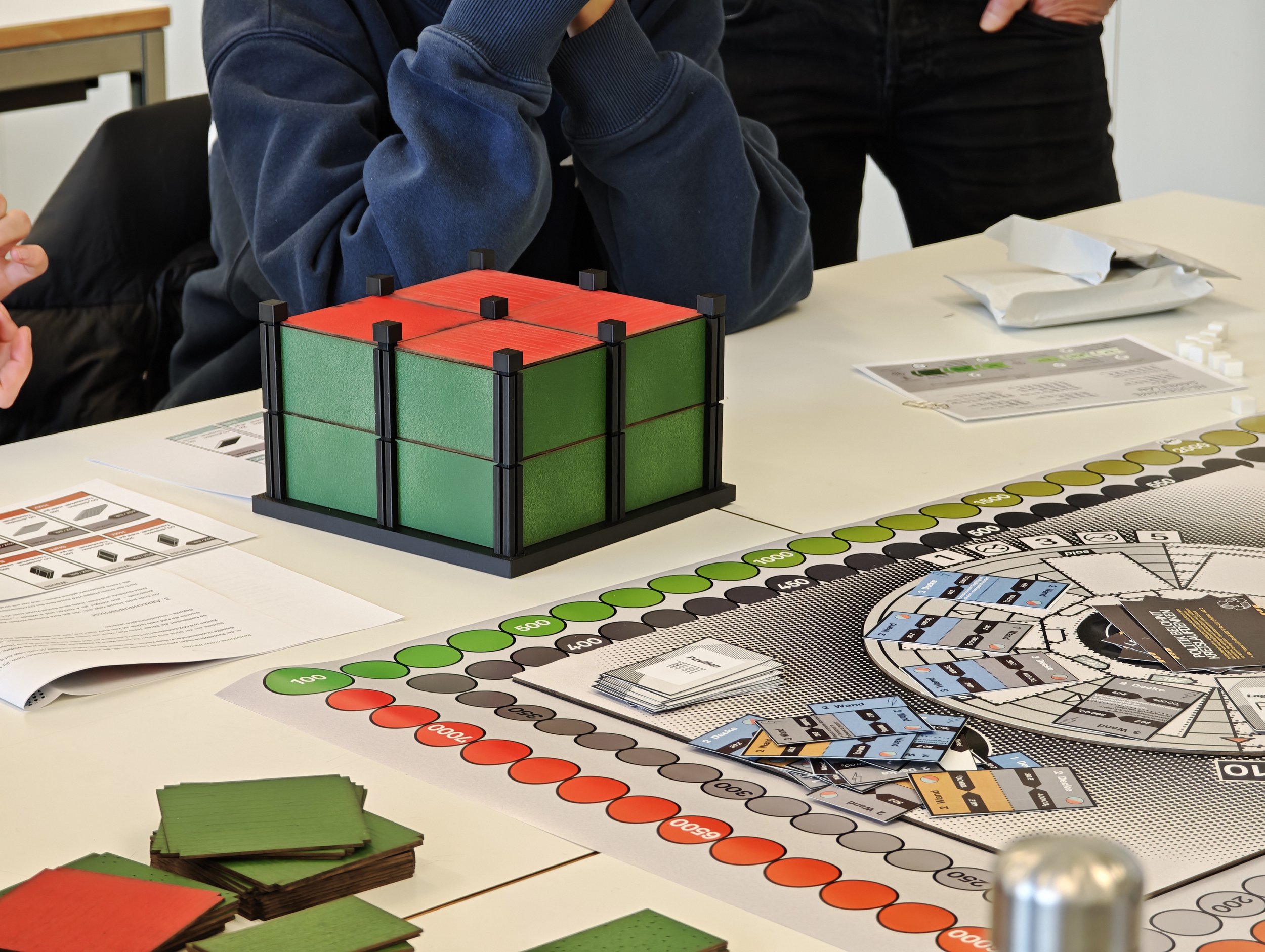 Tisch mit Brettspielen, Spielmaterialien und Spielanleitungen, darunter ein gefalteter Spielplan, Karten, Spielchips und Spielstücke, die für ein Gesellschaftsspiel vorbereitet sind.